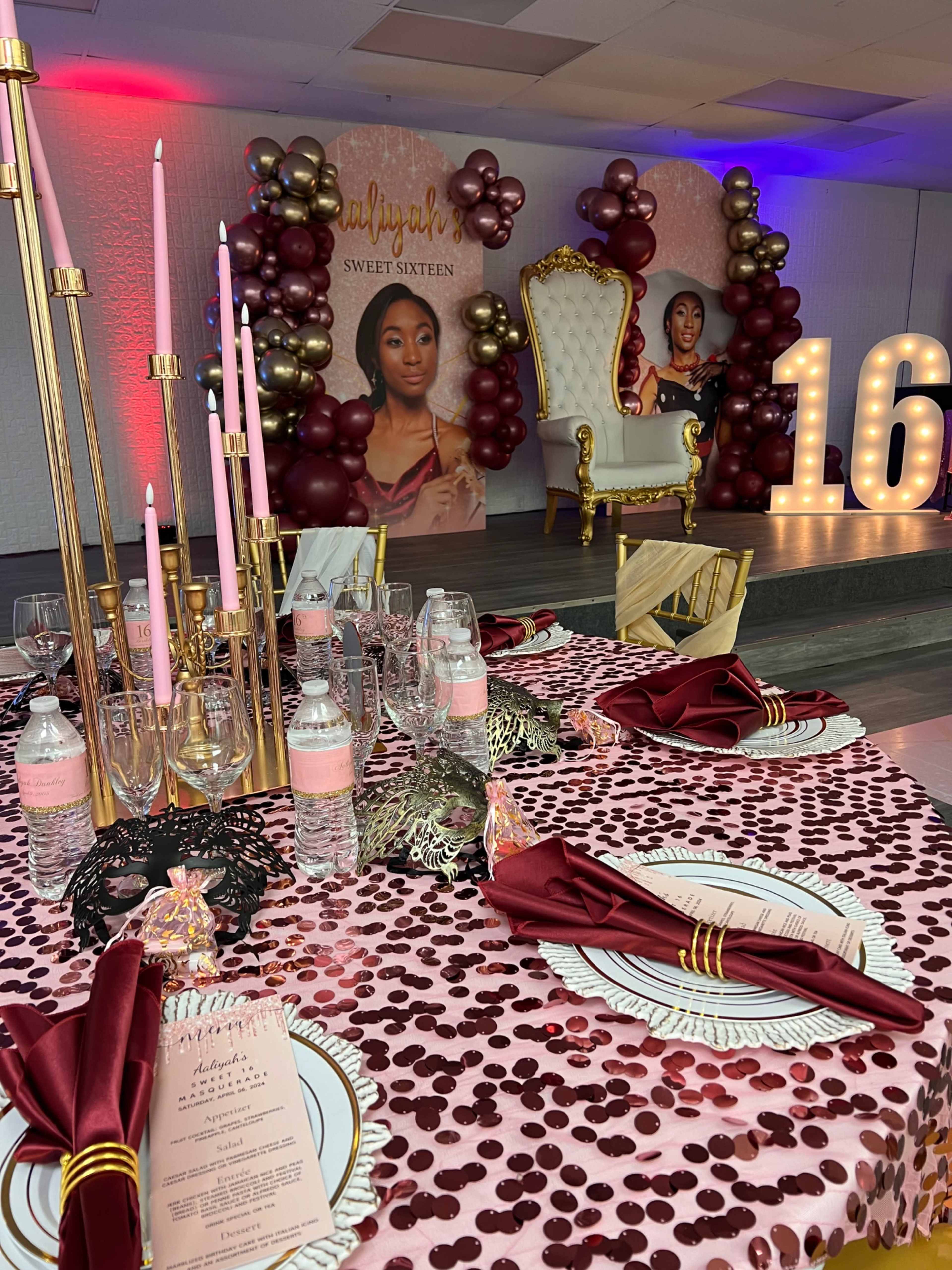 The scene features a decorated indoor space for a sweet sixteen party, complete with a lavish table set with a sequined tablecloth, elegant dinnerware, and a large backdrop displaying a portrait of a young woman alongside a golden throne and a "16" sign made of balloons.