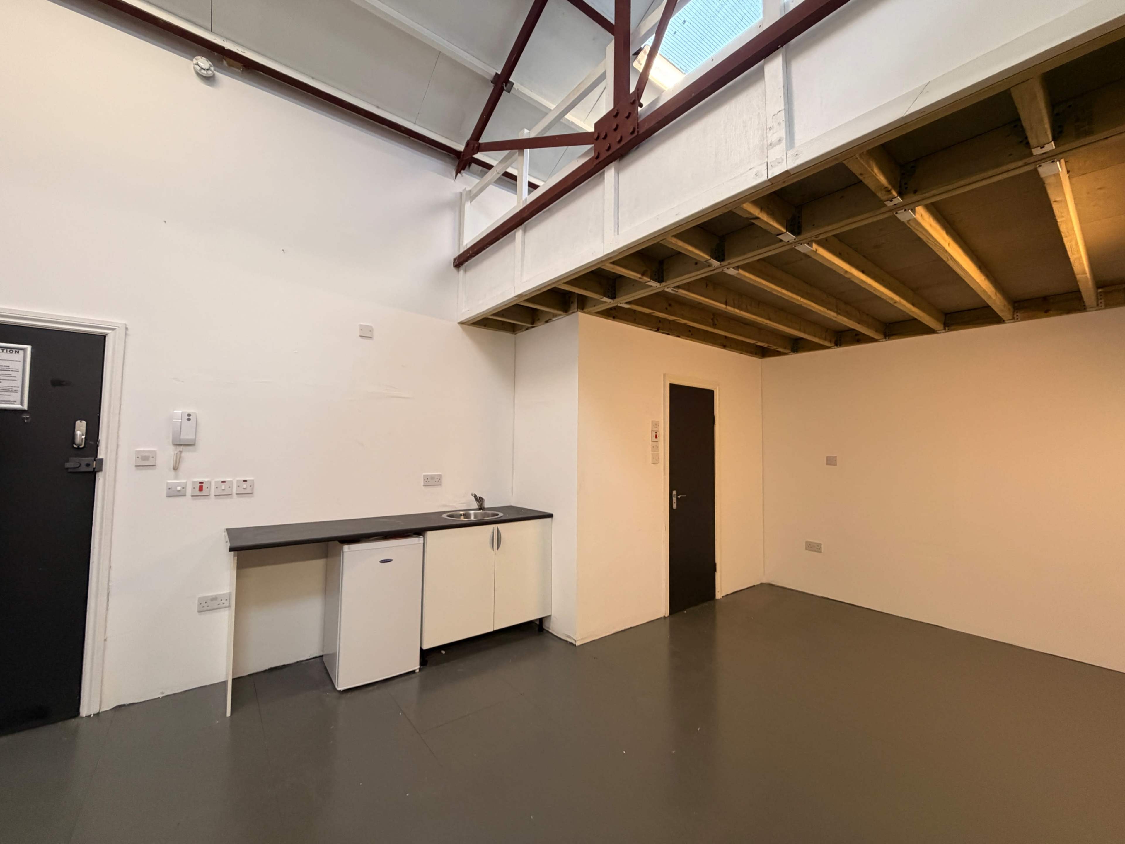 The image shows a spacious room with a counter and sink along one wall, a door on the opposite side, and exposed ceiling beams above.