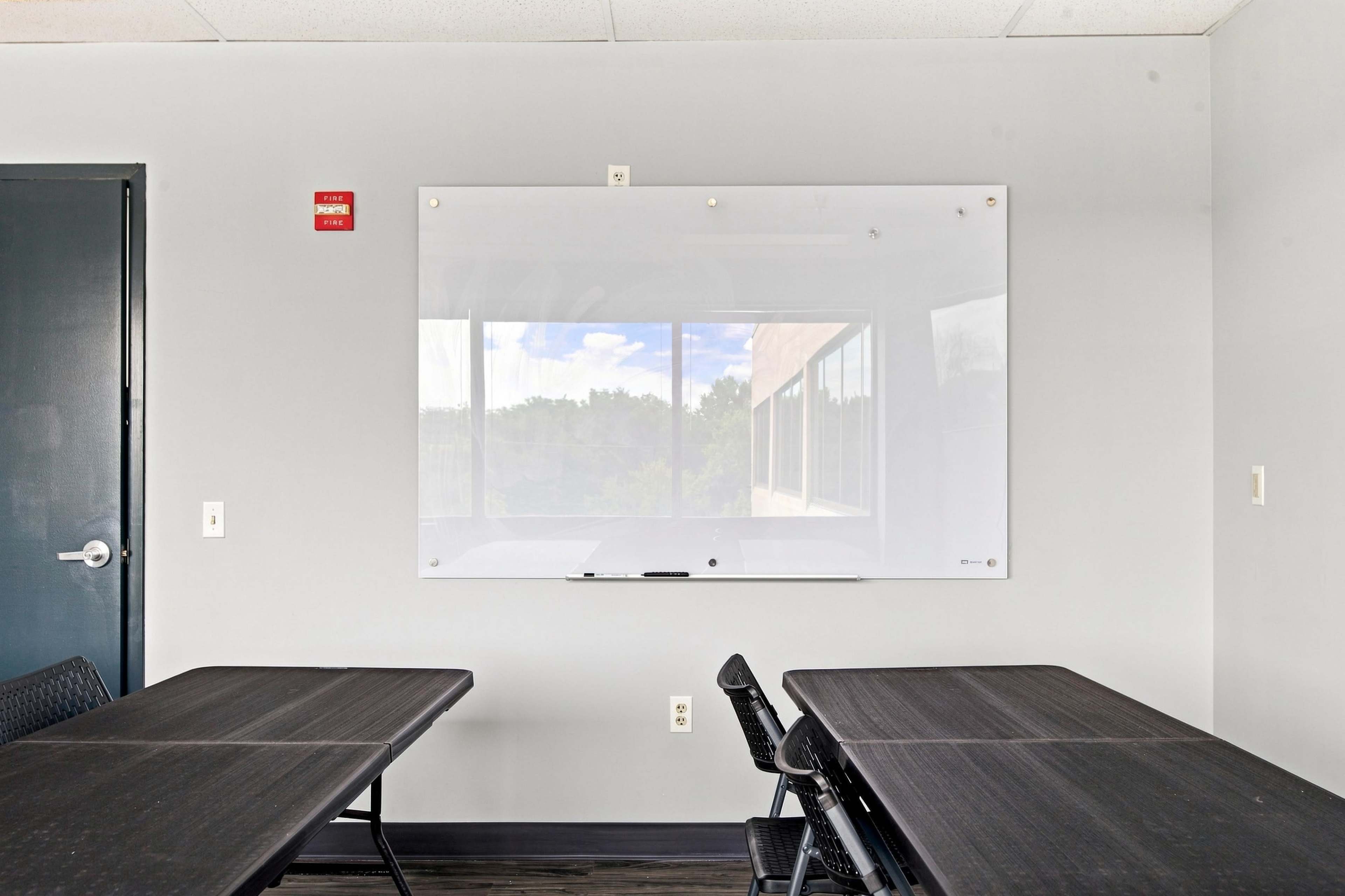 The image shows a conference room with two black tables and a whiteboard mounted on the wall, alongside a large window overlooking greenery.