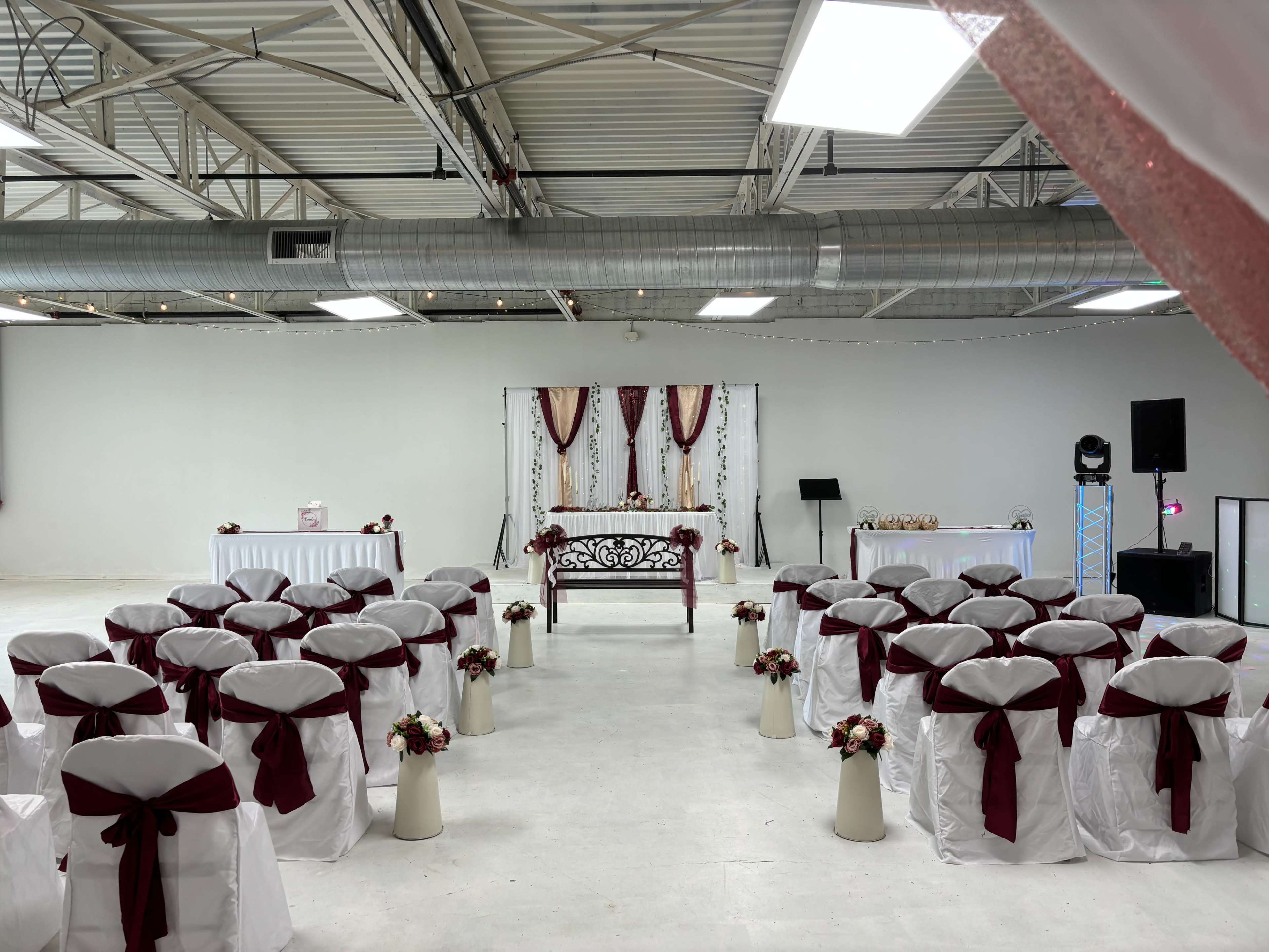 A decorated event space features rows of white and maroon chairs facing a central altar draped with fabric, surrounded by floral arrangements.