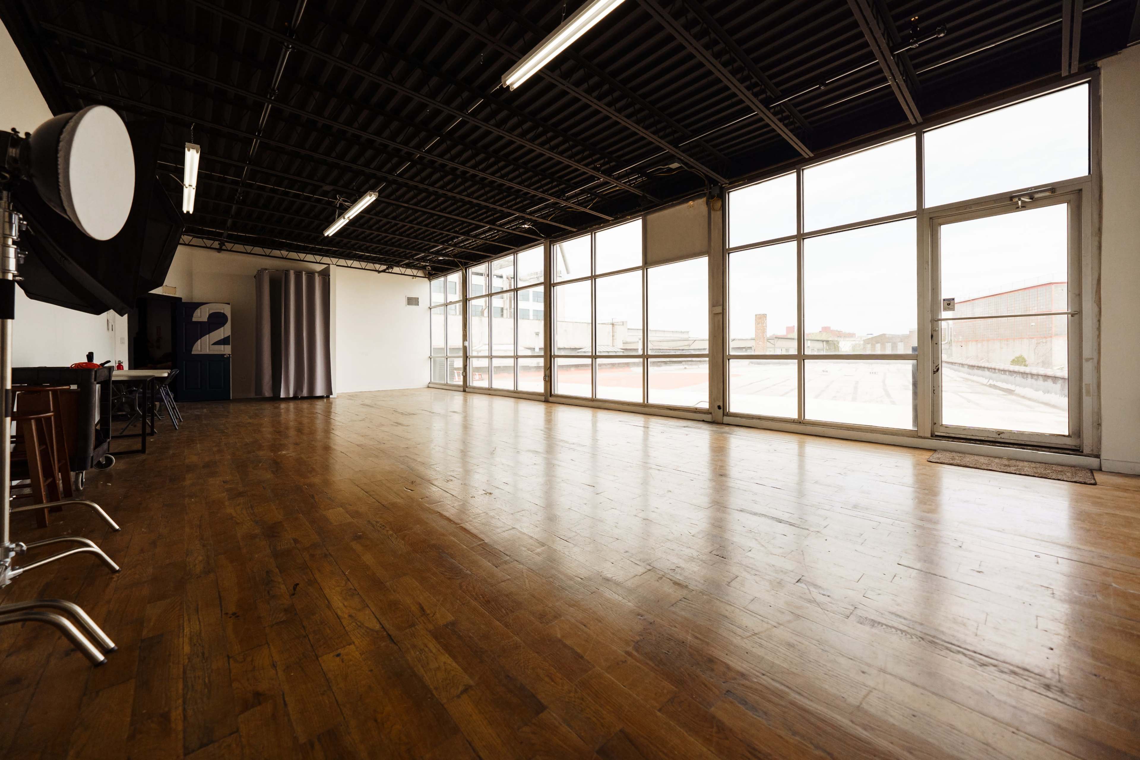 The image shows a spacious, empty studio with wooden flooring and large windows allowing natural light to enter.