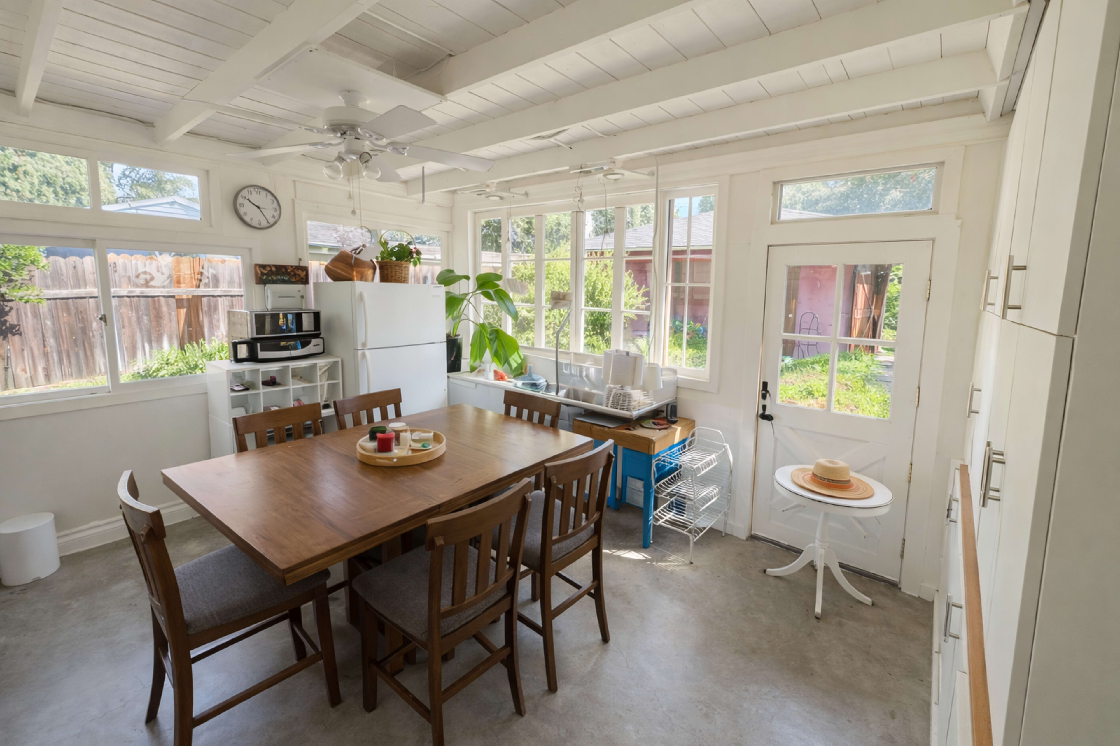 The image shows a bright kitchen with a wooden dining table, chairs, and large windows that let in natural light.