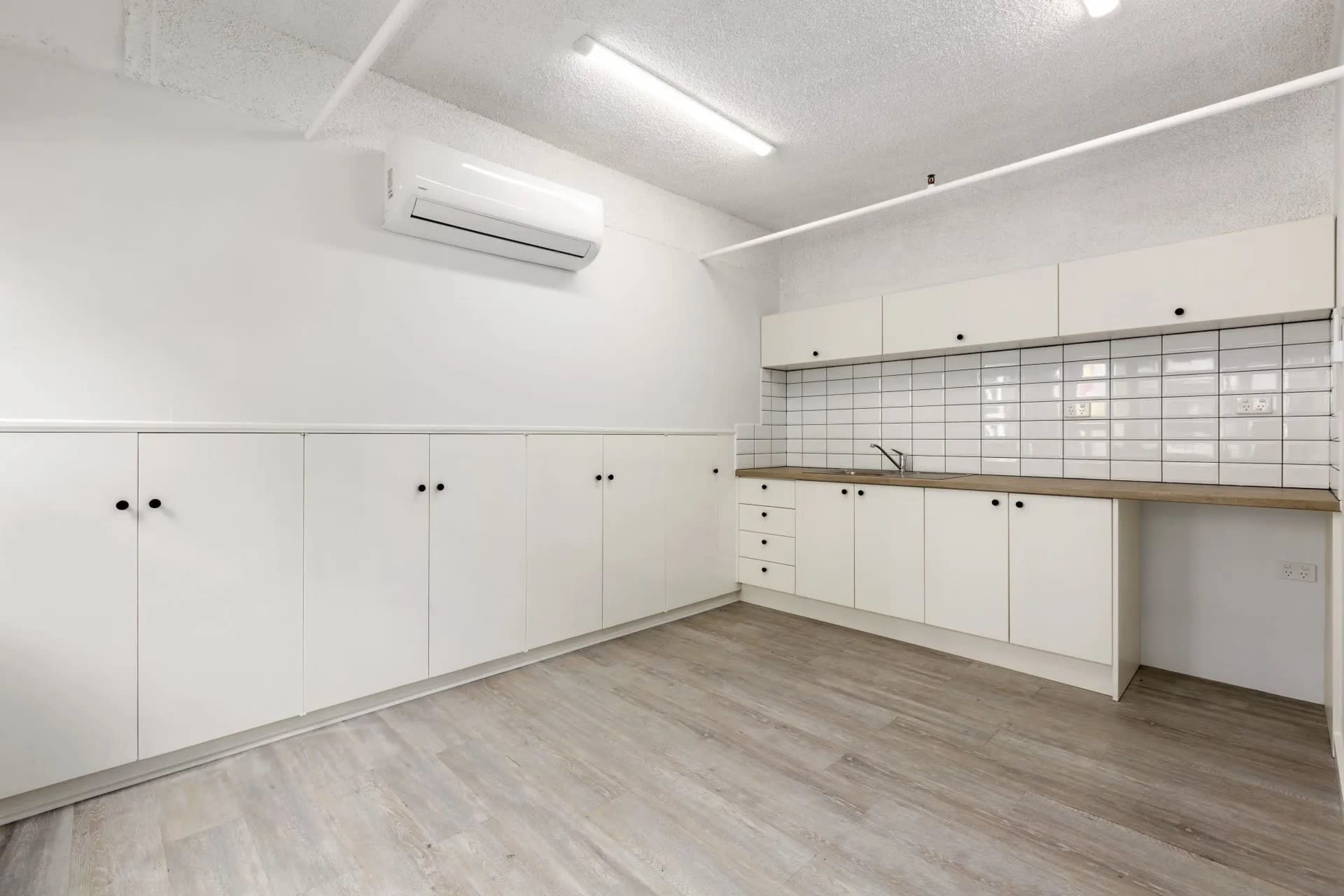 The image shows a minimalist kitchen with white cabinets, a wooden countertop, and a tiled backsplash, featuring an air conditioning unit on the ceiling.