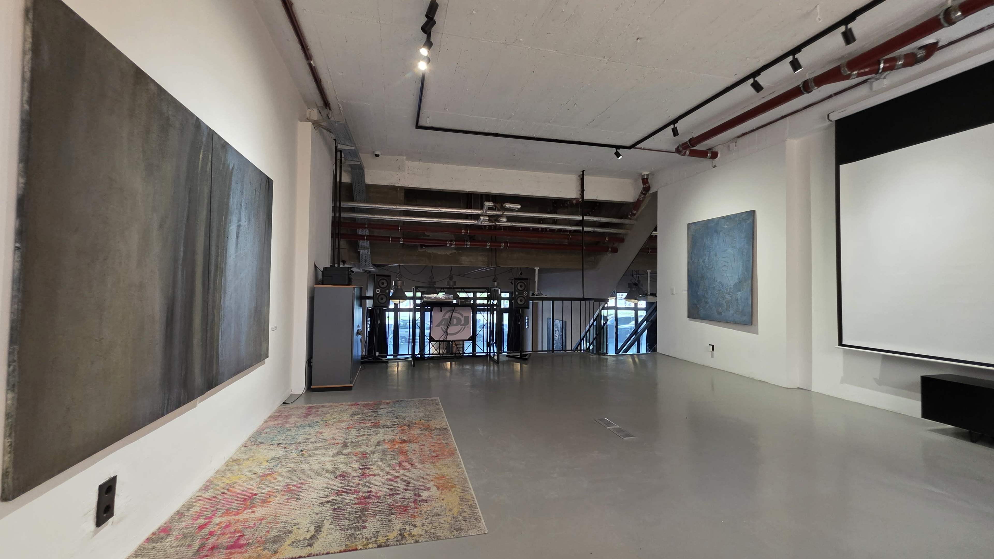 The image shows an art gallery interior with two large paintings on the walls, an area rug on the floor, and a projected screen against one wall.
