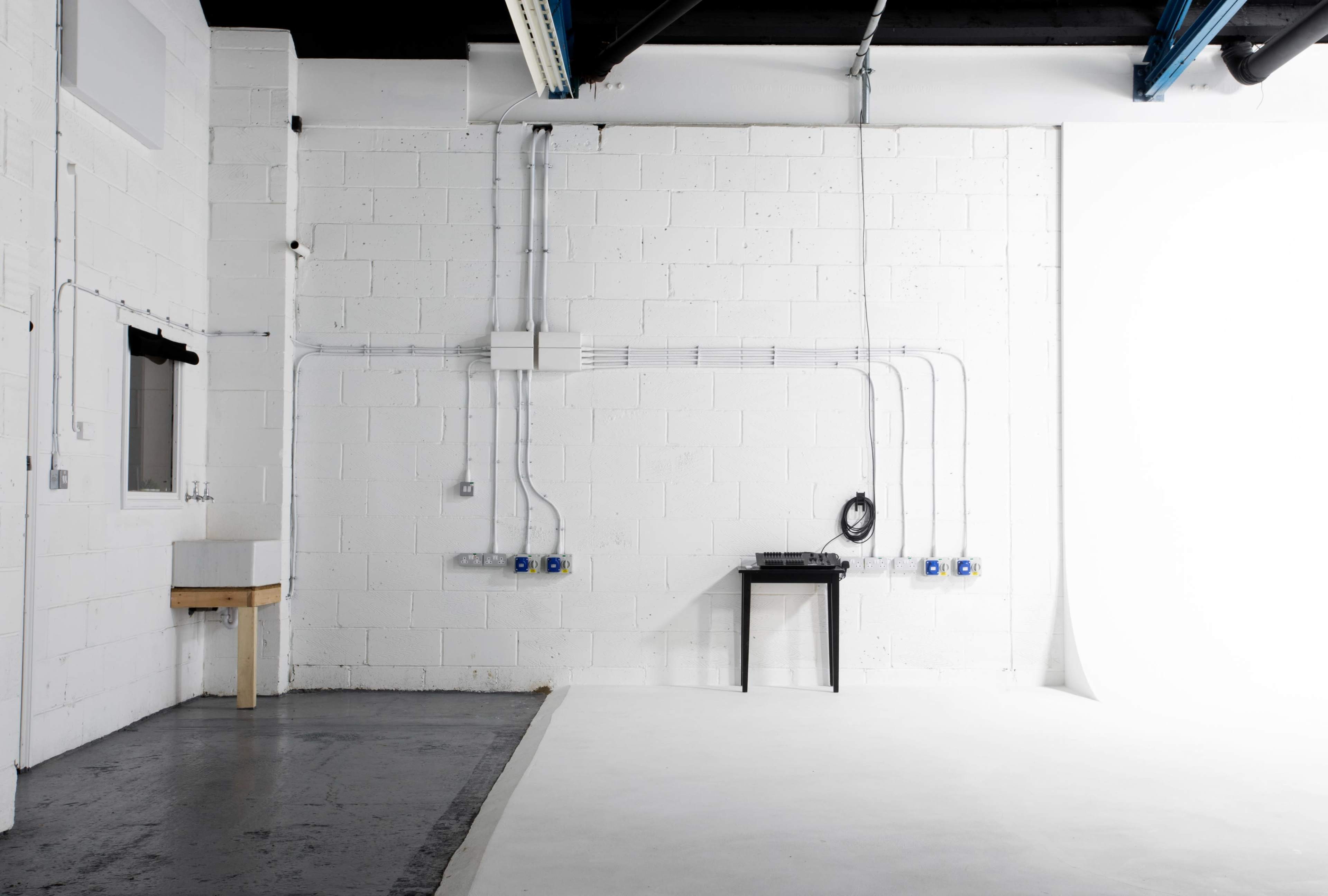 A sparsely furnished room with white walls, concrete flooring, and various electrical outlets along the wall, along with a small table and a sink in one corner.