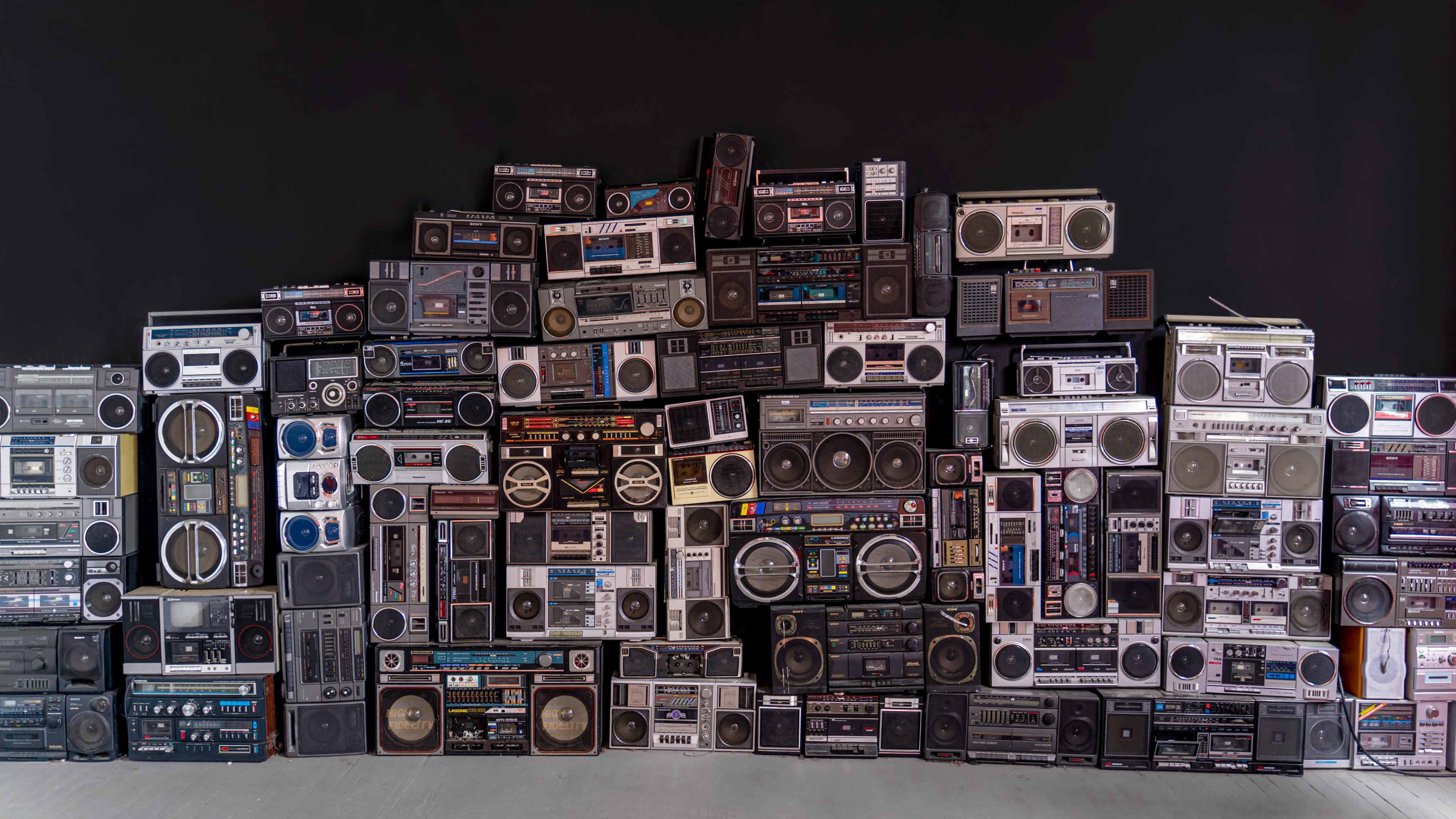 A large collection of vintage boomboxes is arranged in a stacked formation against a black background.