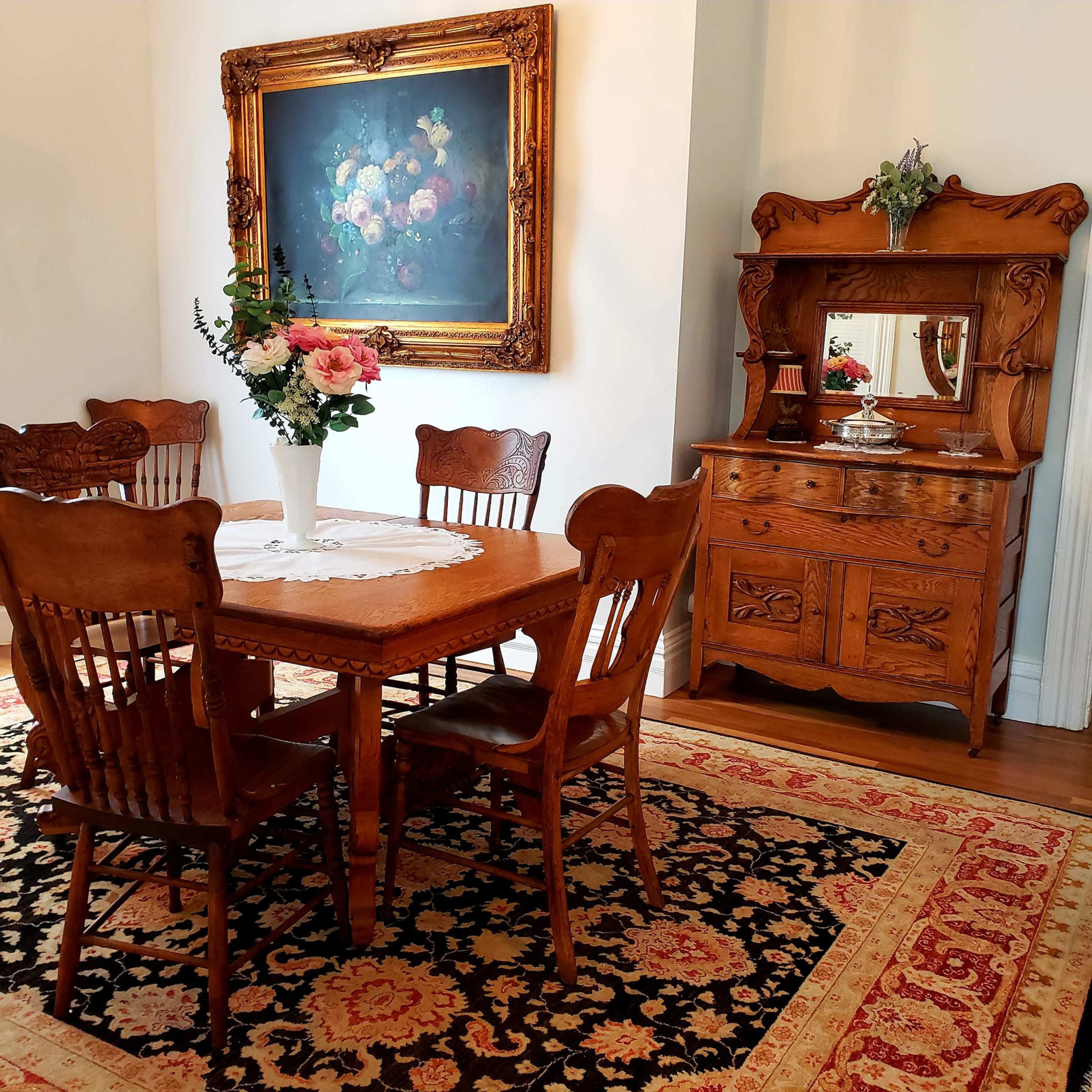 A wooden dining table surrounded by chairs is set in a room with a vintage hutch and a large floral painting on the wall.