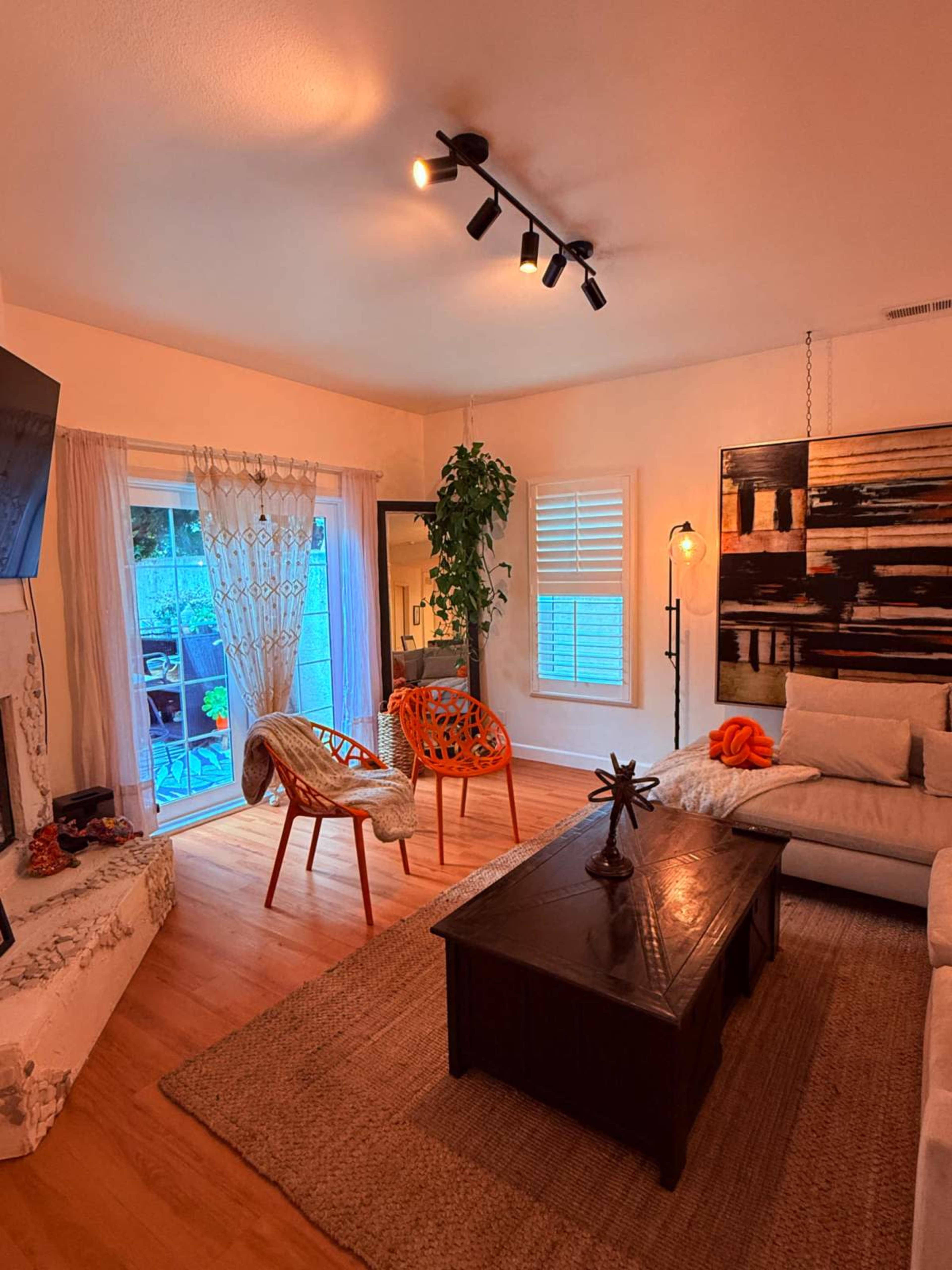 The image shows a cozy living room with a light-colored sofa, a wooden coffee table, and two red accent chairs near a glass door that opens to an outdoor area.