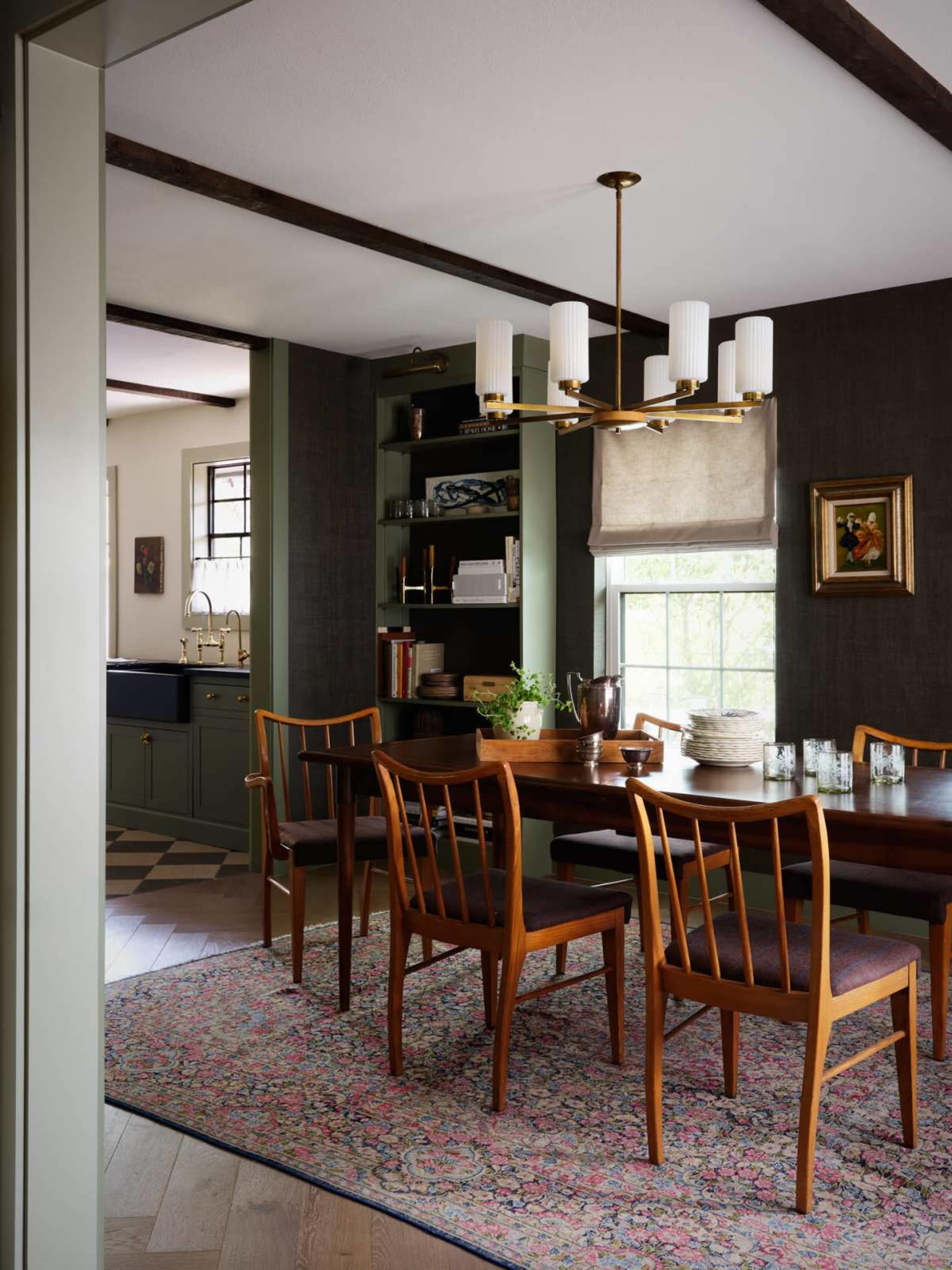 A dining room features a wooden table surrounded by six chairs, with a chandelier overhead and a patterned rug beneath.