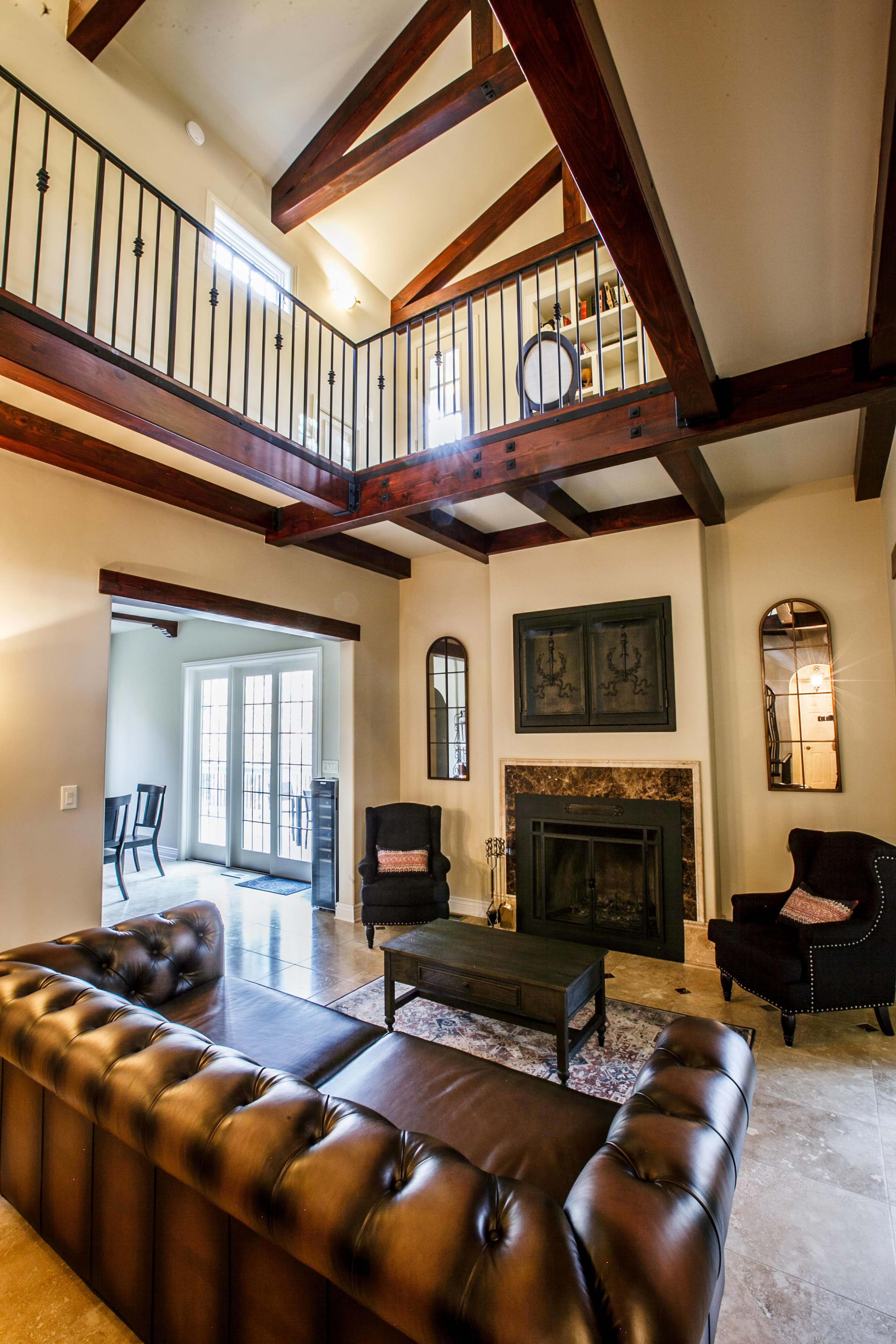 A spacious living room featuring a brown leather couch, a coffee table, a fireplace, and wooden beams overhead, with access to a balcony above.