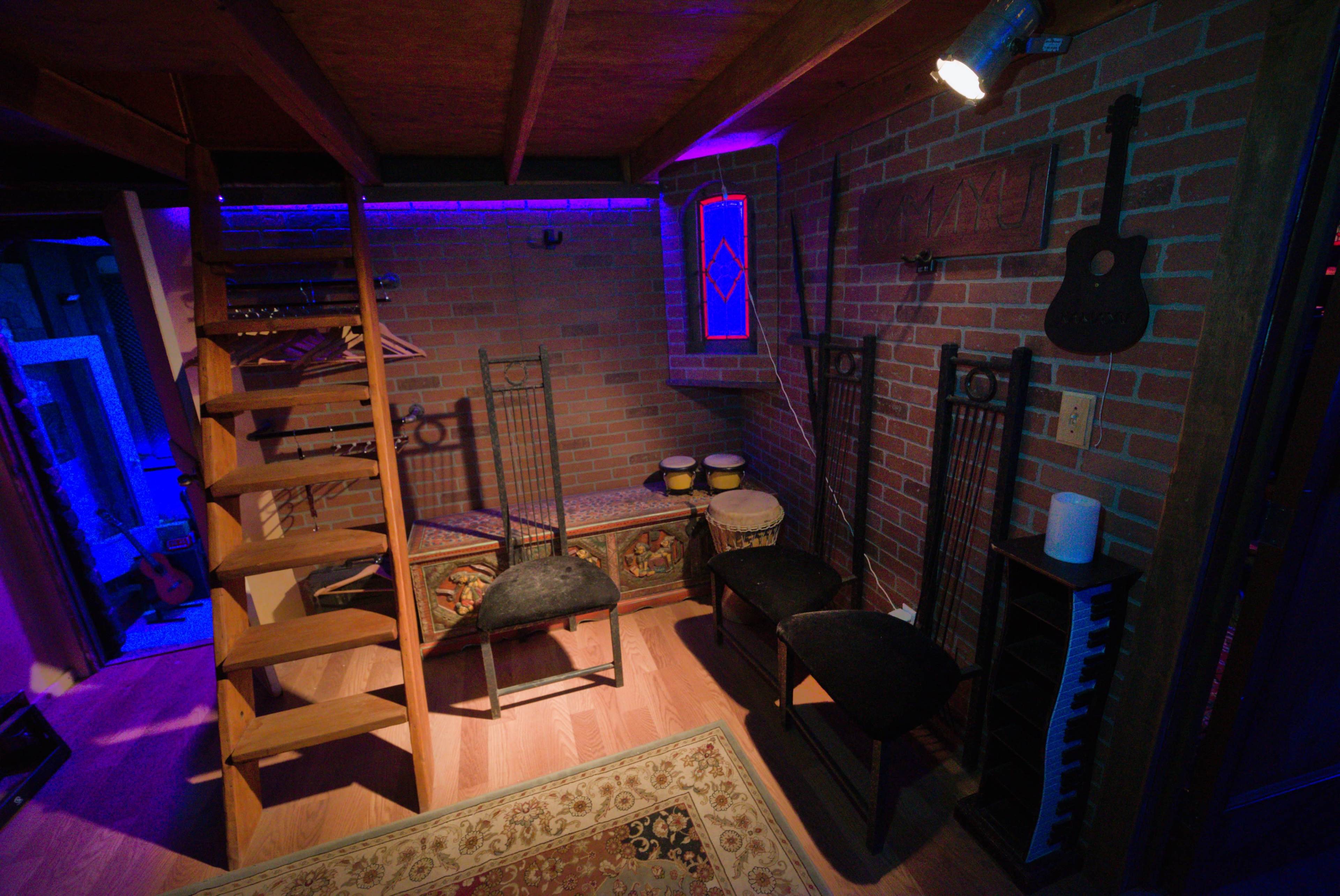 The image shows a cozy basement space with a wooden staircase, three black chairs, a decorative storage bench, conga drums, and a soft rug under warm lighting.