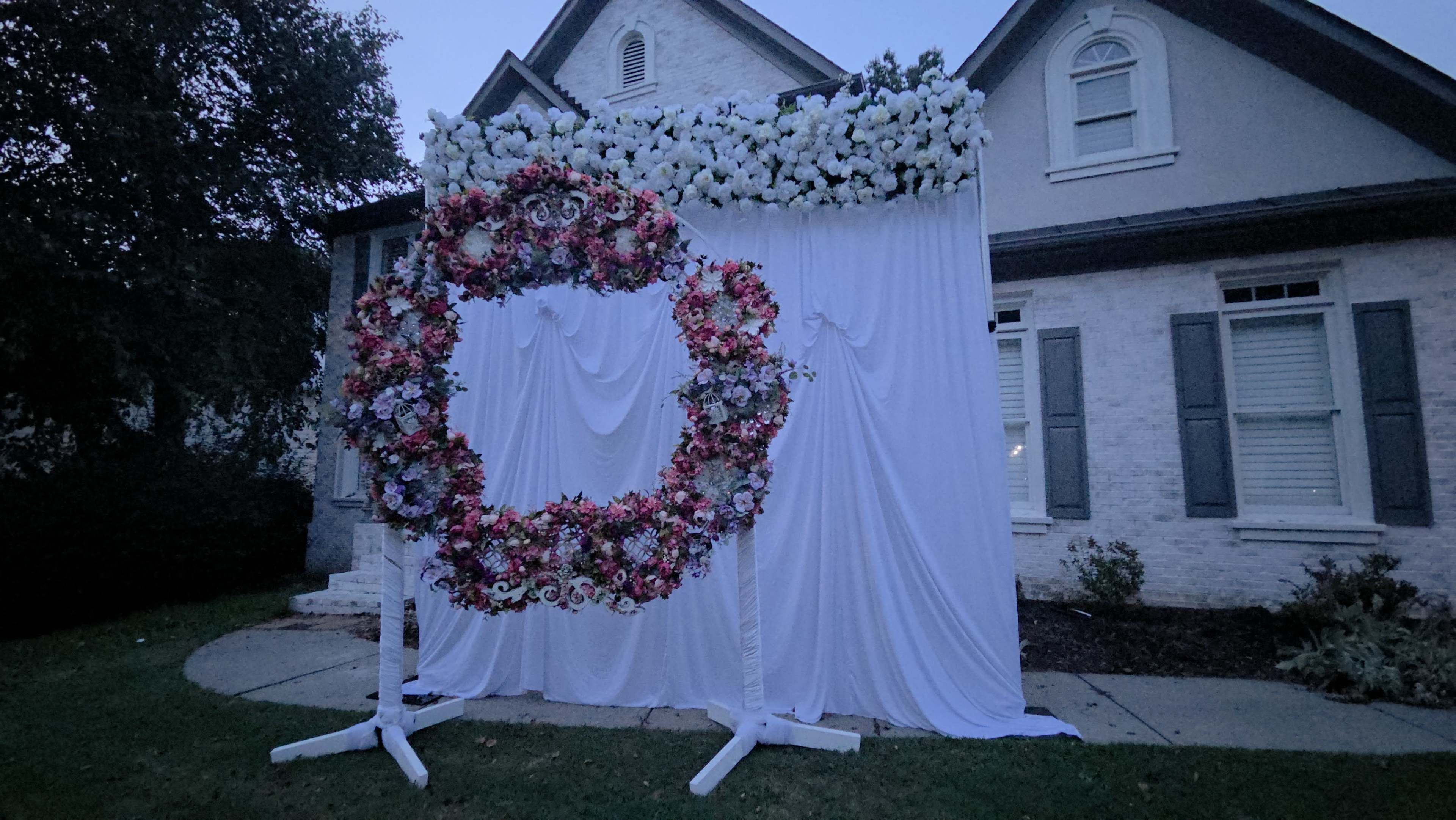 A large floral wreath adorned with various colored flowers is mounted on a white backdrop draped in a residential yard.