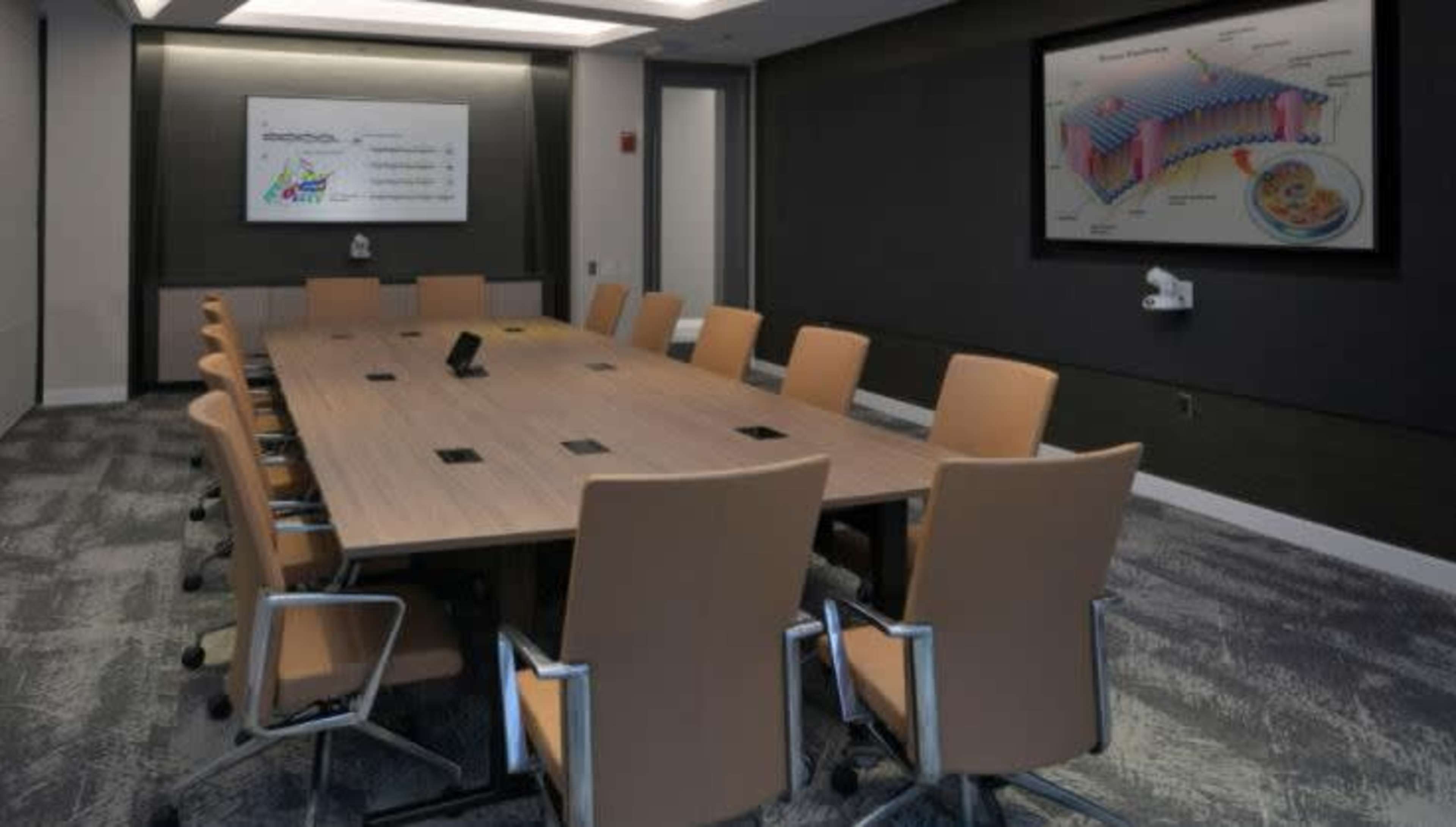 A large conference room features a long wooden table surrounded by ergonomic chairs, with a wall display showing a diagram.