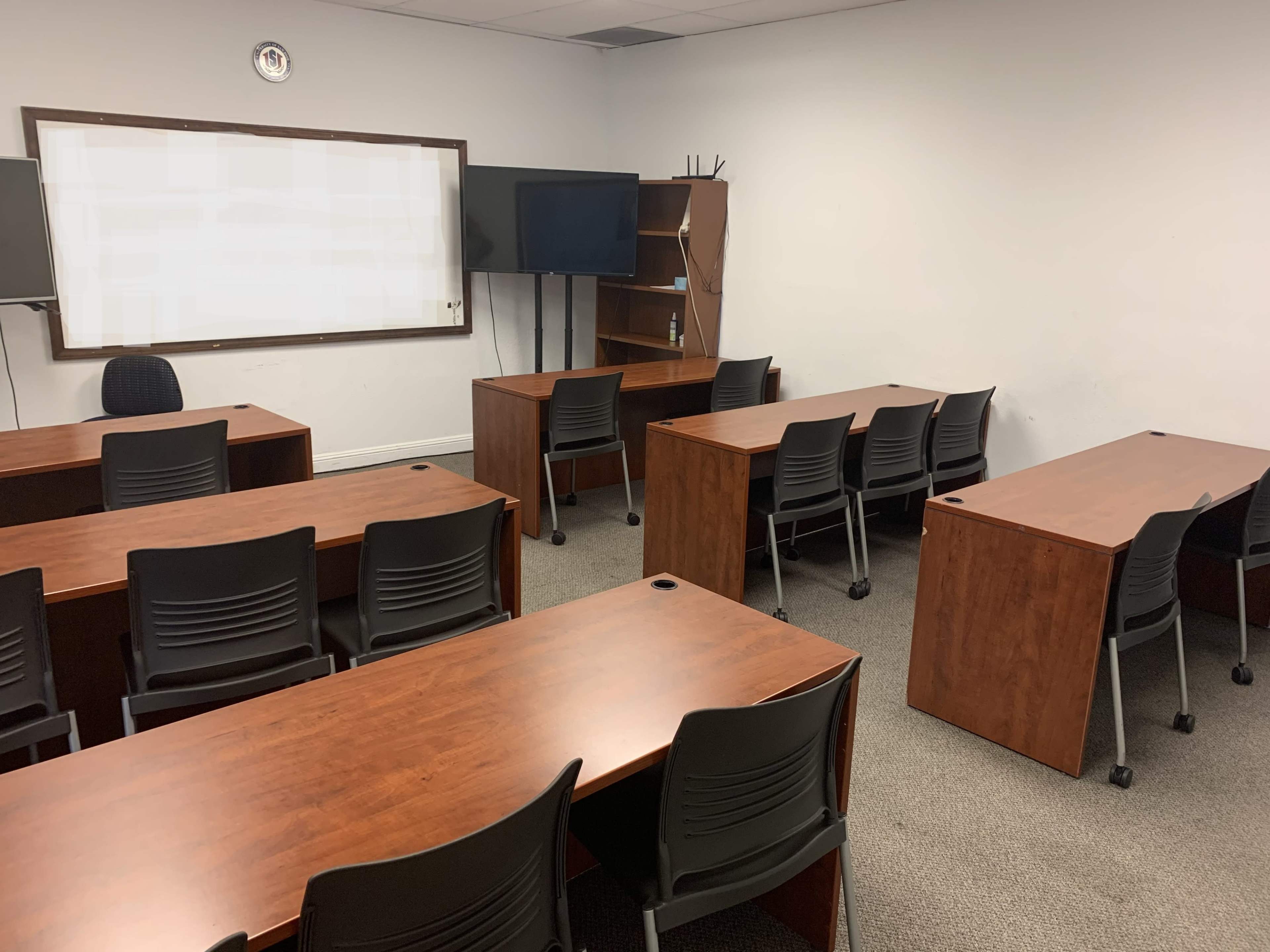 An empty classroom with wooden tables and black chairs arranged in several rows, along with a whiteboard and a television on the wall.