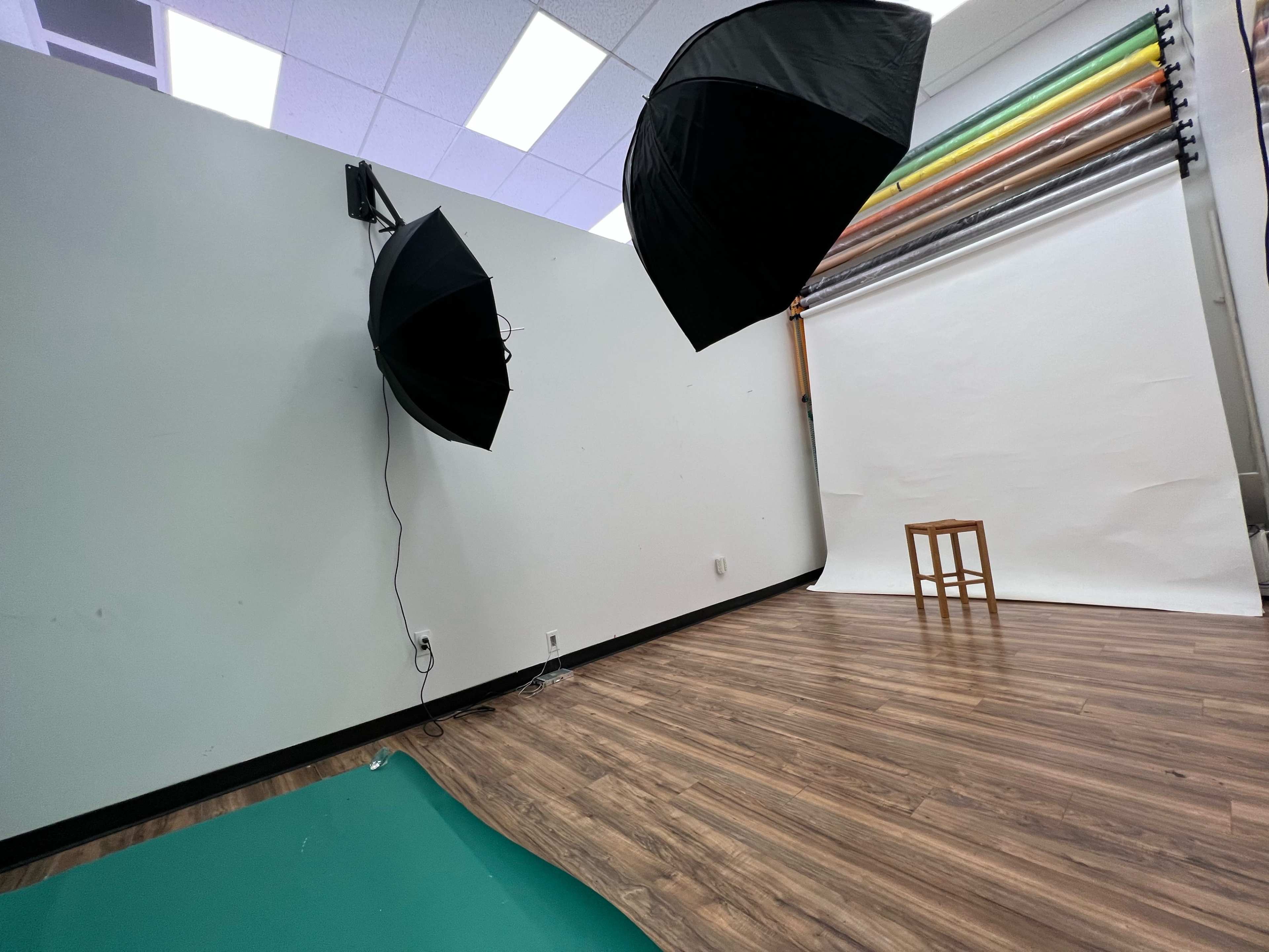 A photography studio setup with two softbox lights, a white backdrop, and a wooden stool on a hardwood floor.