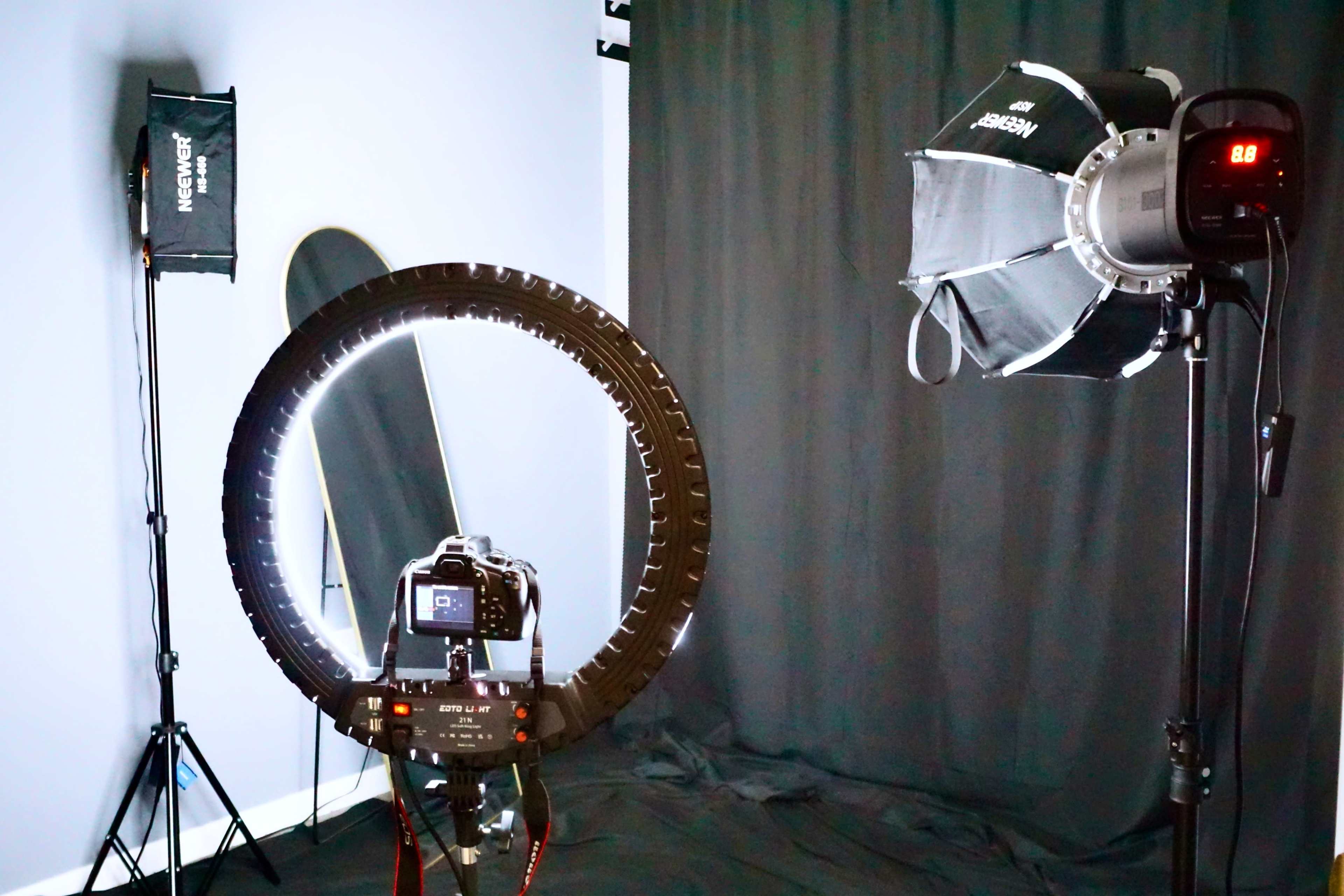 The image shows a photography studio setup featuring a camera mounted on a tripod, a ring light, and a softbox light against a dark backdrop.