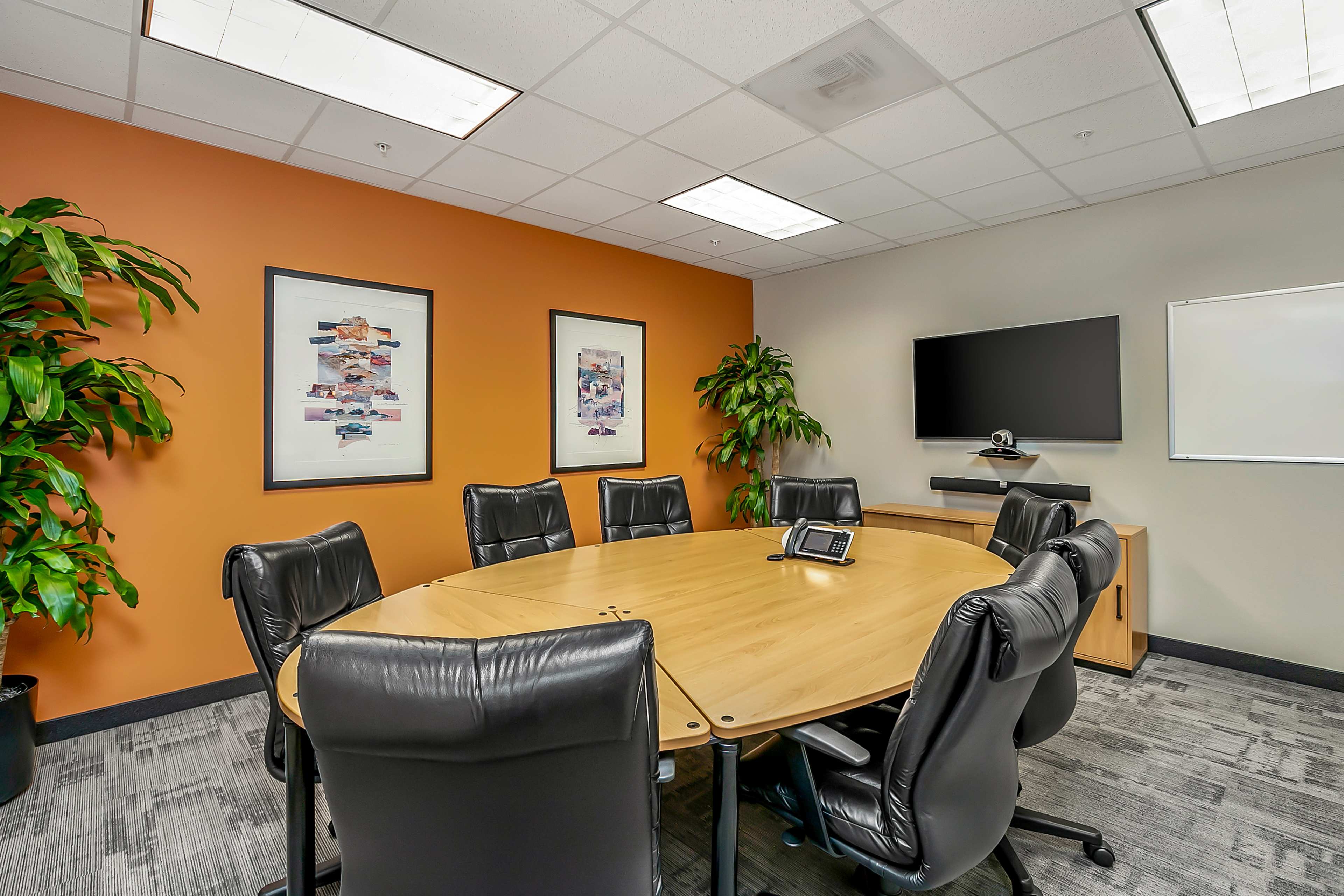 A conference room features a large round wooden table surrounded by black leather chairs, with art on the walls, plants in the corners, and a wall-mounted TV.