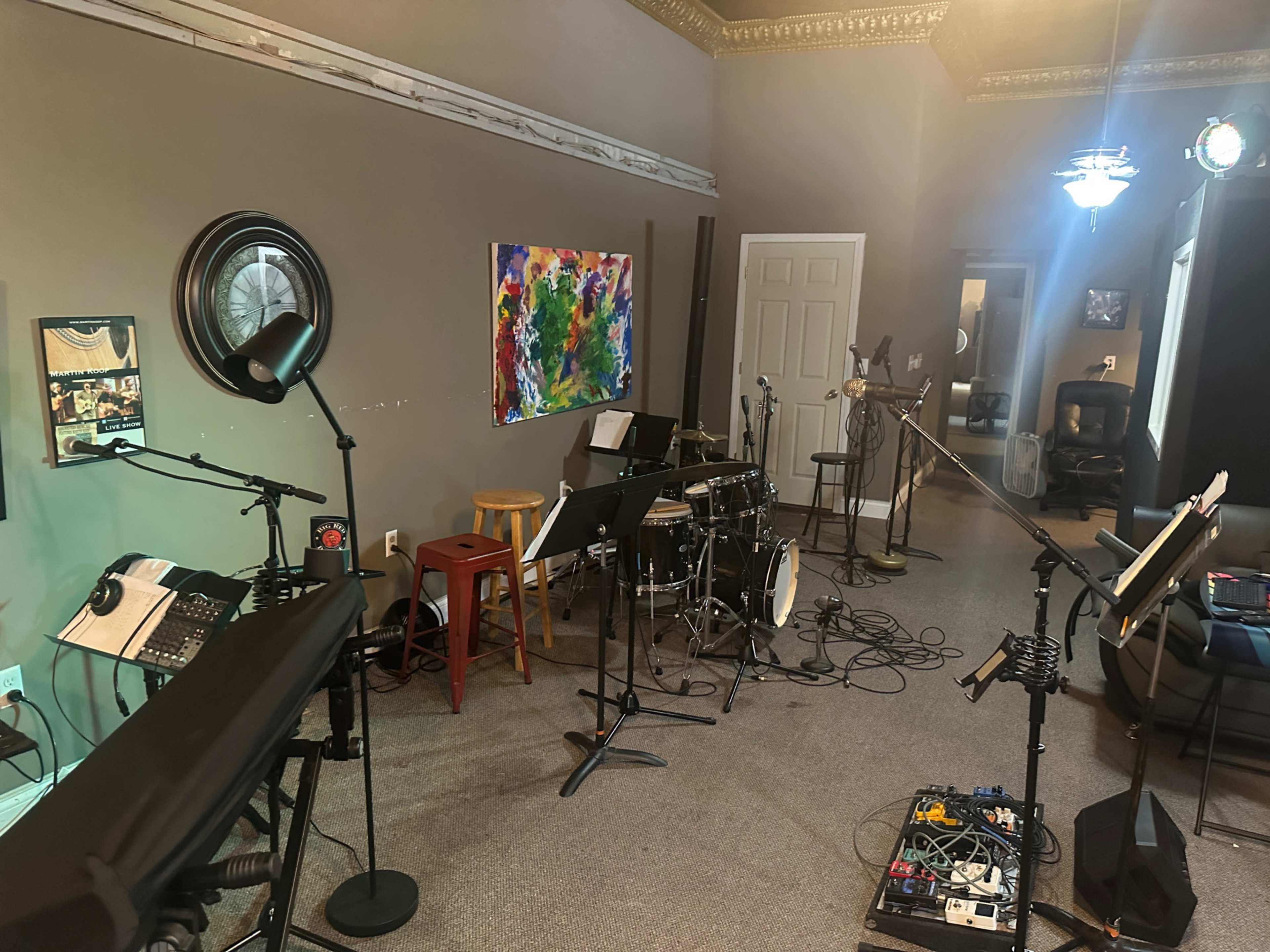 The image shows a music studio with various instruments, including a drum set and keyboards, along with microphones and music stands arranged throughout the space.