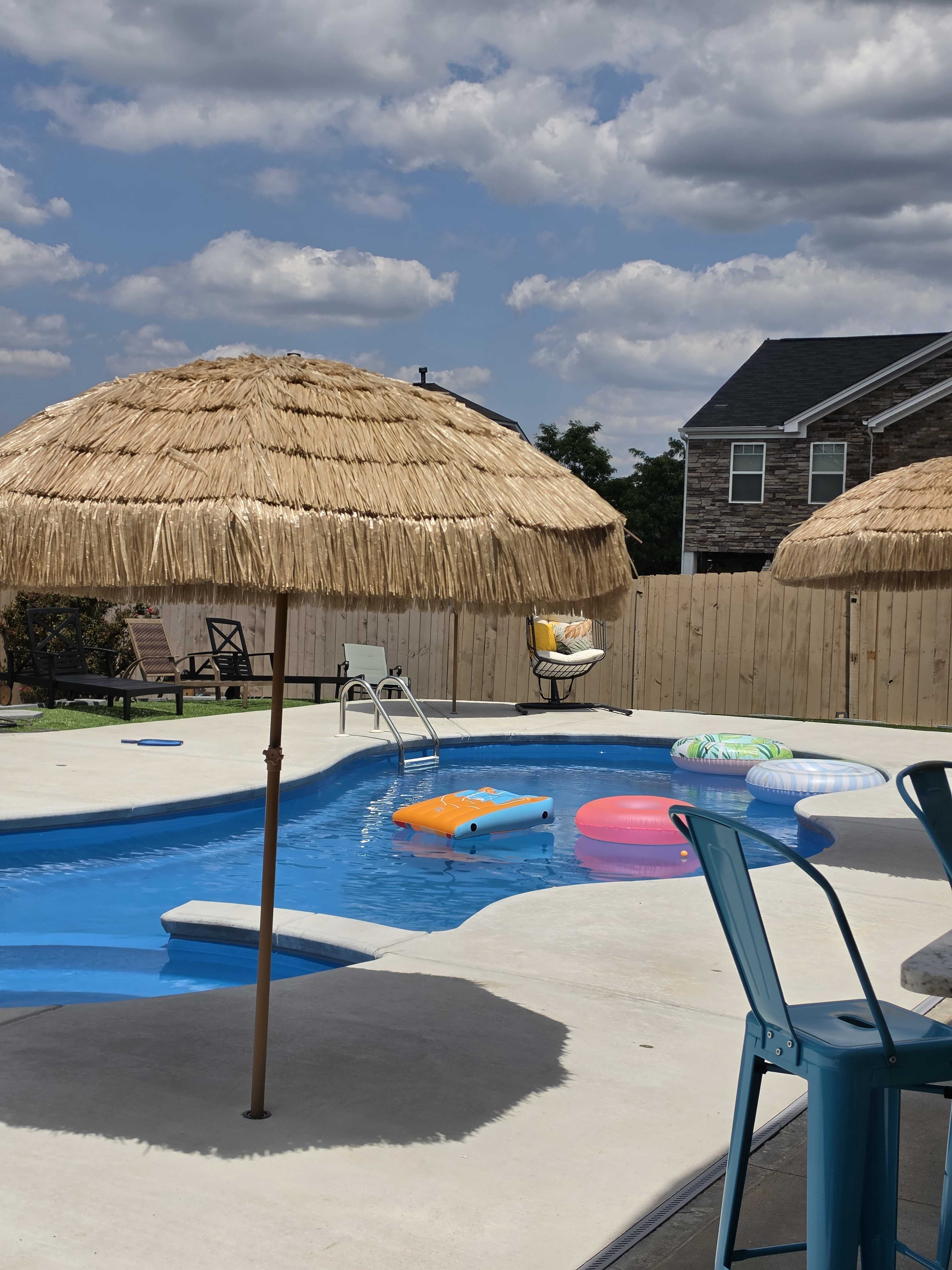 The image shows a swimming pool surrounded by straw umbrellas and colorful inflatable floats on a sunny day.