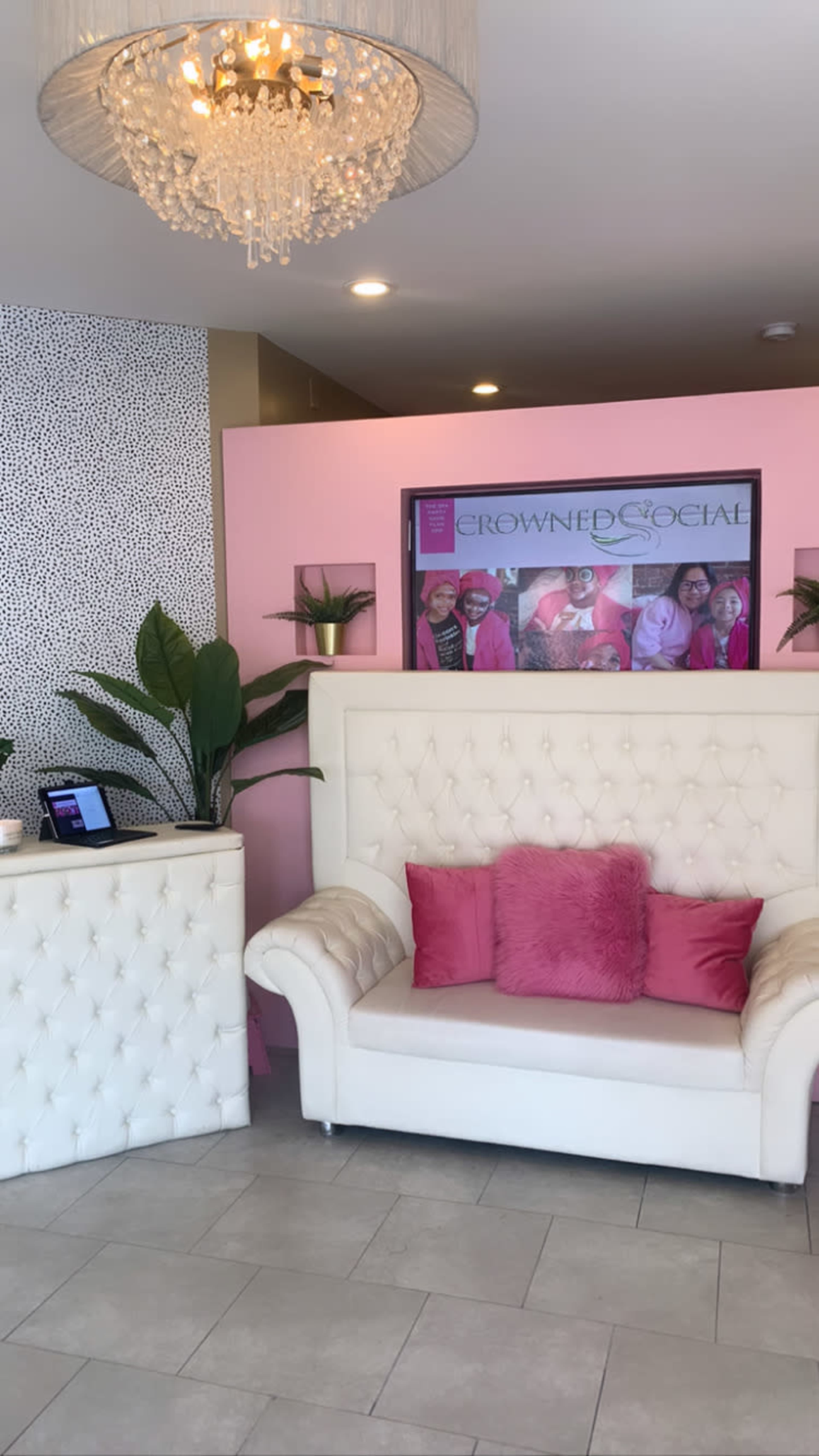 A modern lounge area with a white tufted sofa adorned with pink cushions, a sleek reception desk, and a framed promotional poster on the wall.