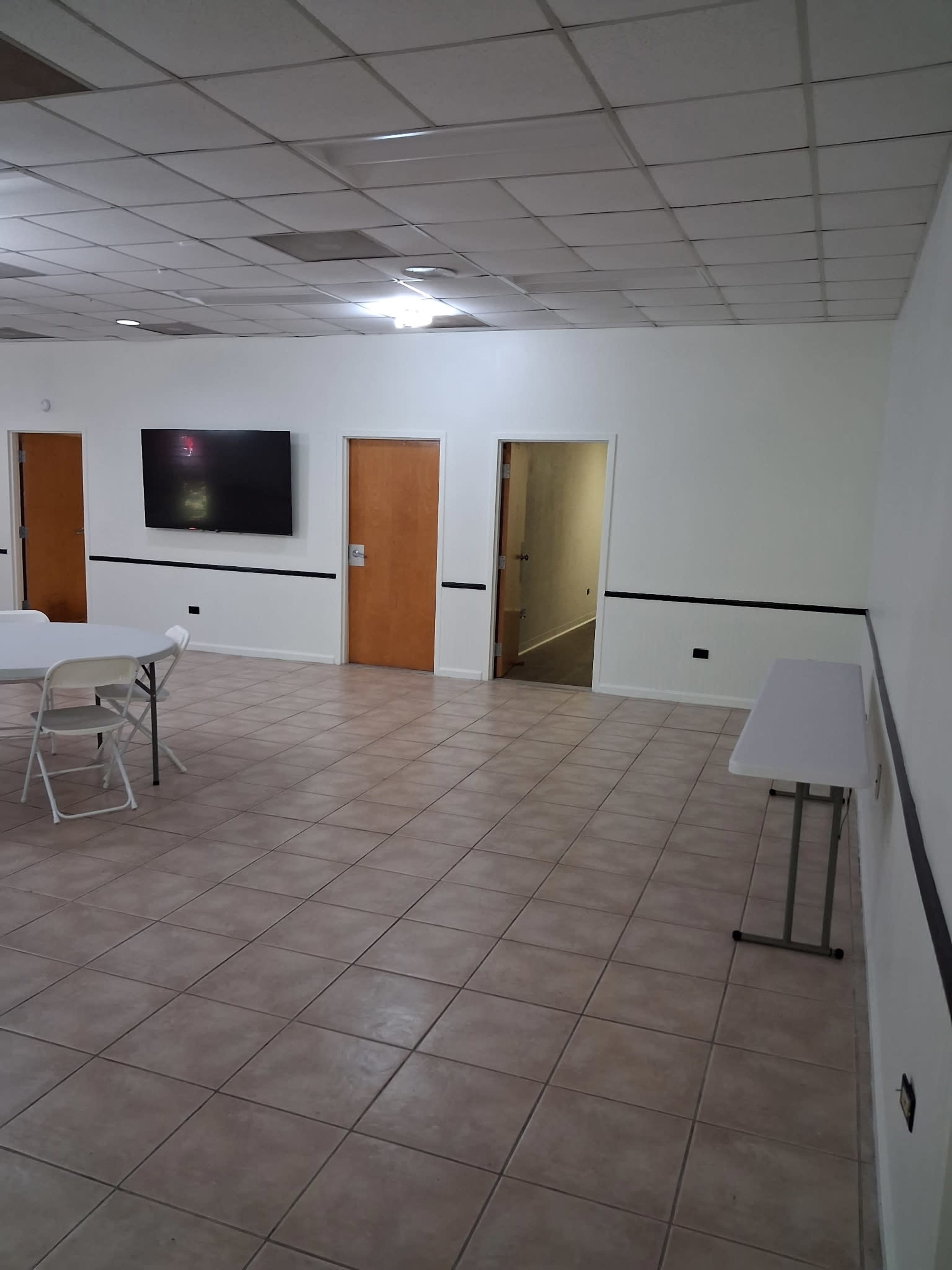 The image shows a spacious, well-lit room with tiled flooring, folding tables, and doors leading to another area.