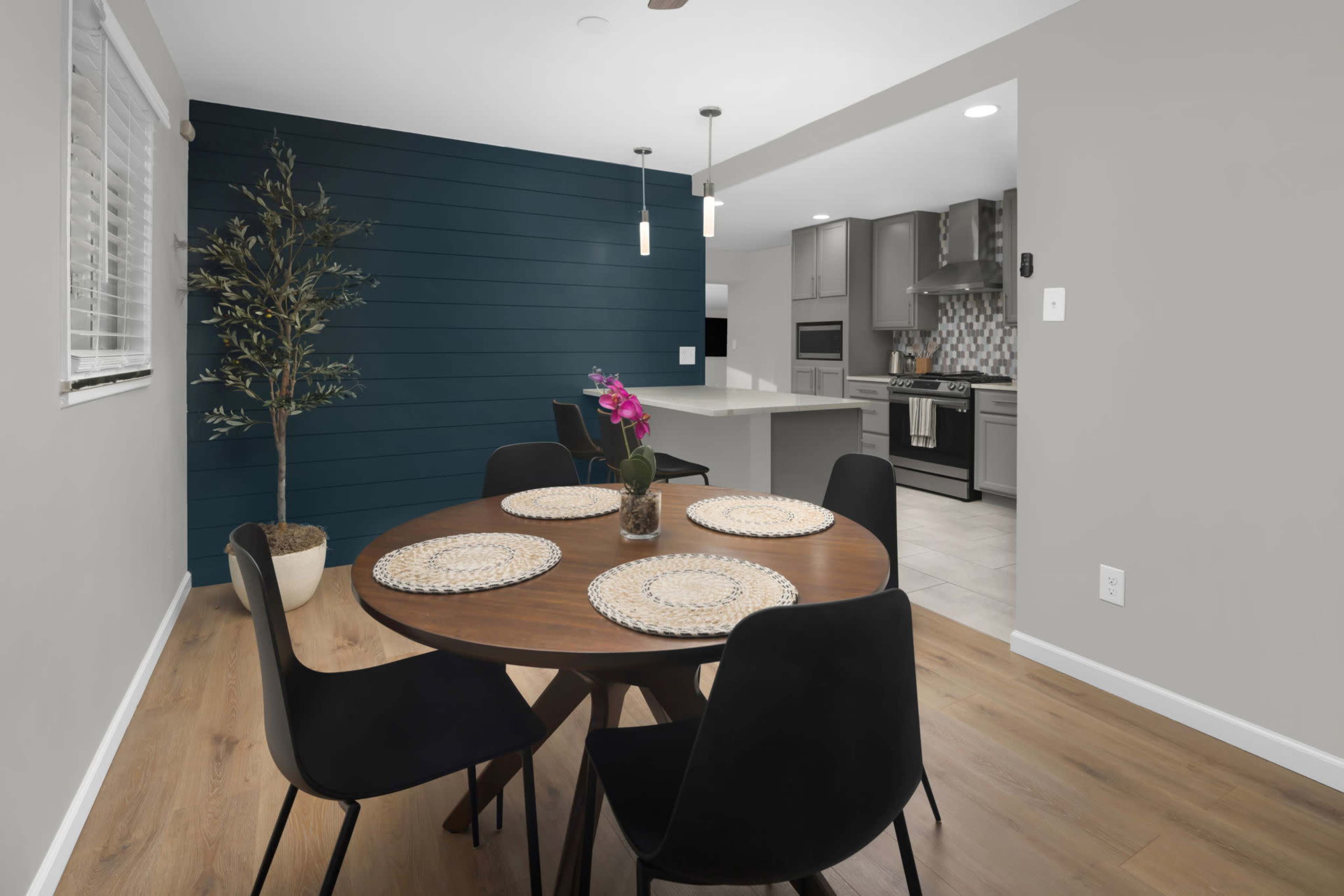 A dining area features a round wooden table with black chairs, a potted plant, and a backdrop of a blue accent wall leading to a modern kitchen.