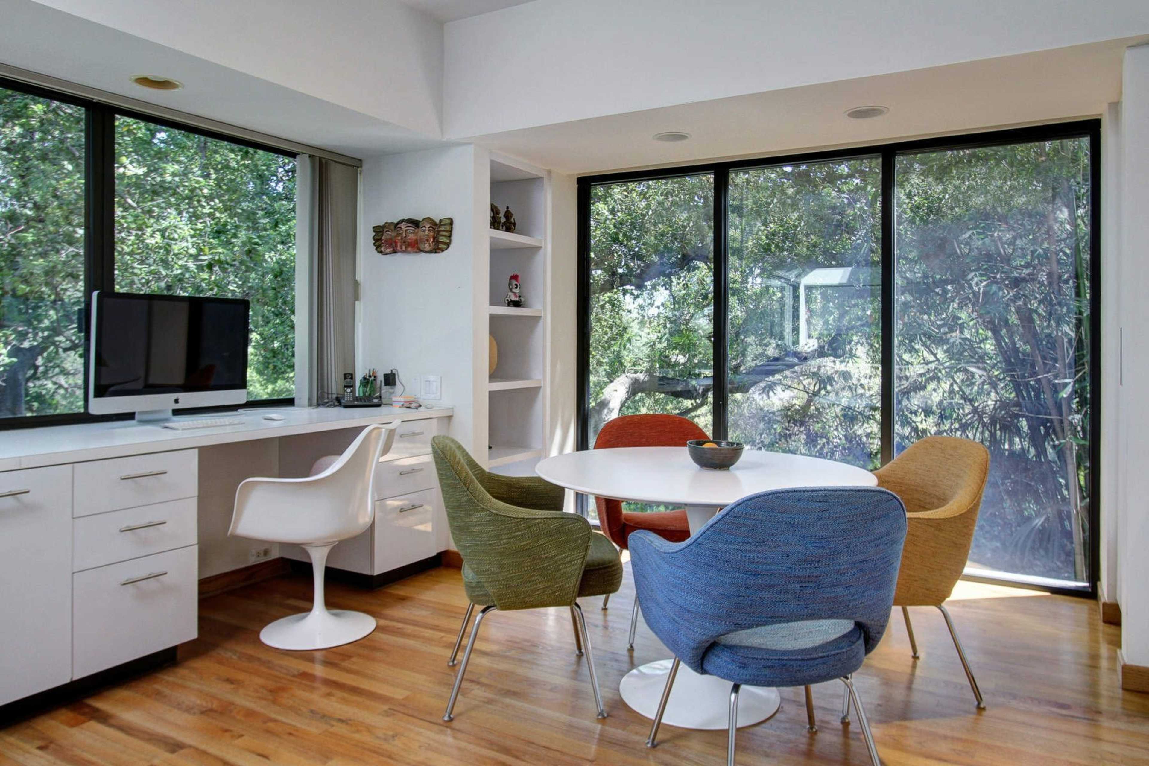 A modern dining area features a round table surrounded by colorful chairs, with a desktop computer and large windows overlooking greenery.