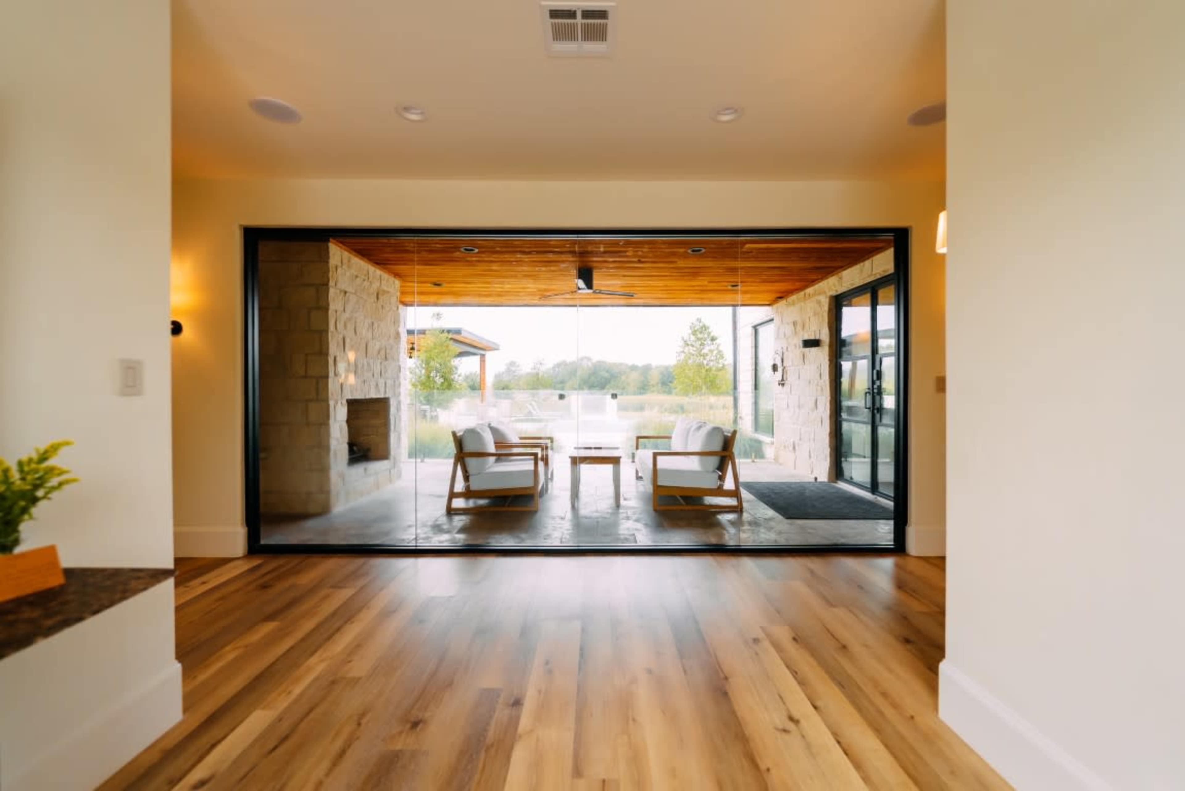 A modern living space opens to an outdoor area through large glass doors, featuring wooden ceilings and a stone fireplace.