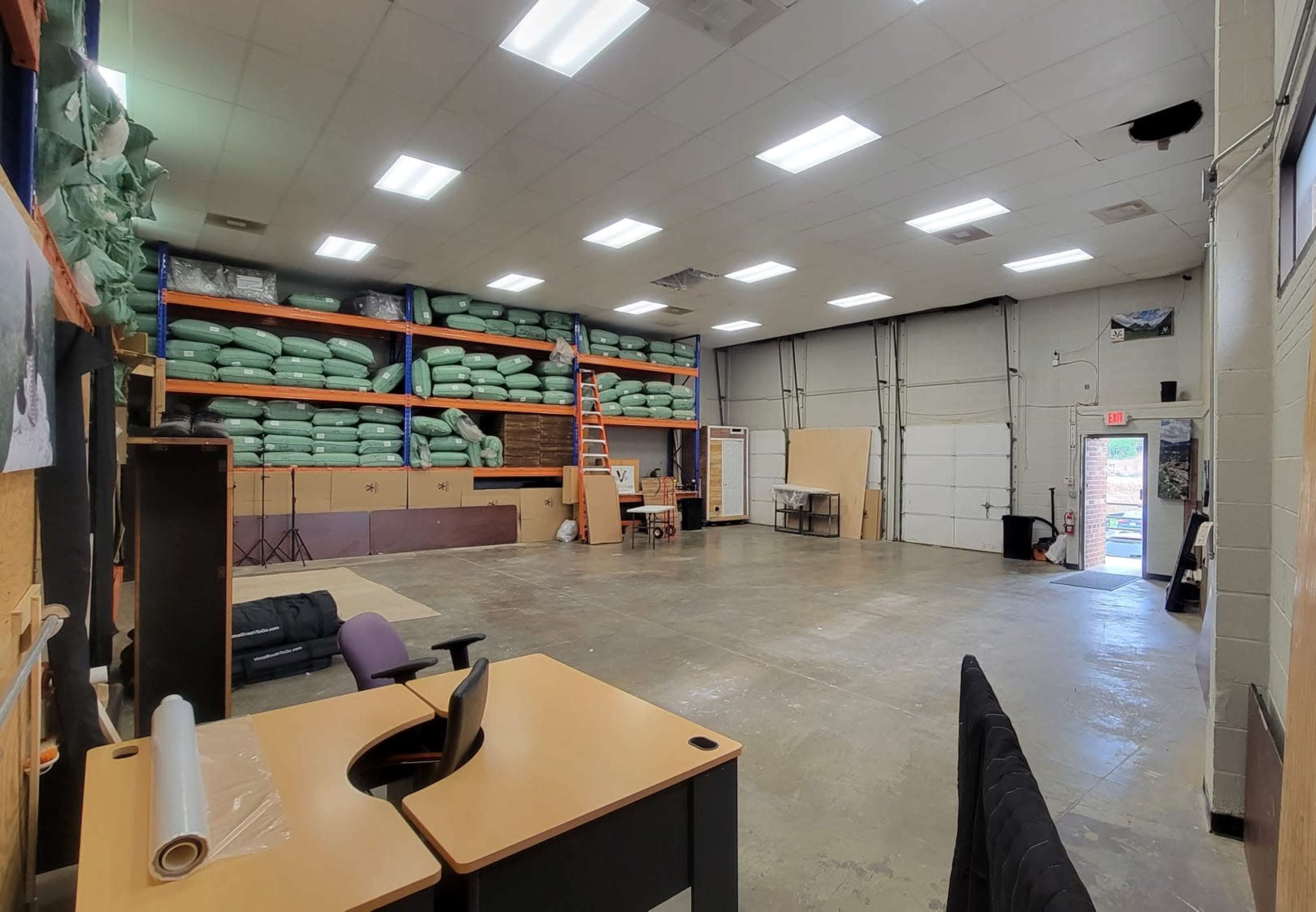 A spacious warehouse interior with green materials stacked on shelves, a ladder leaning against the shelves, and a loading dock visible in the background.