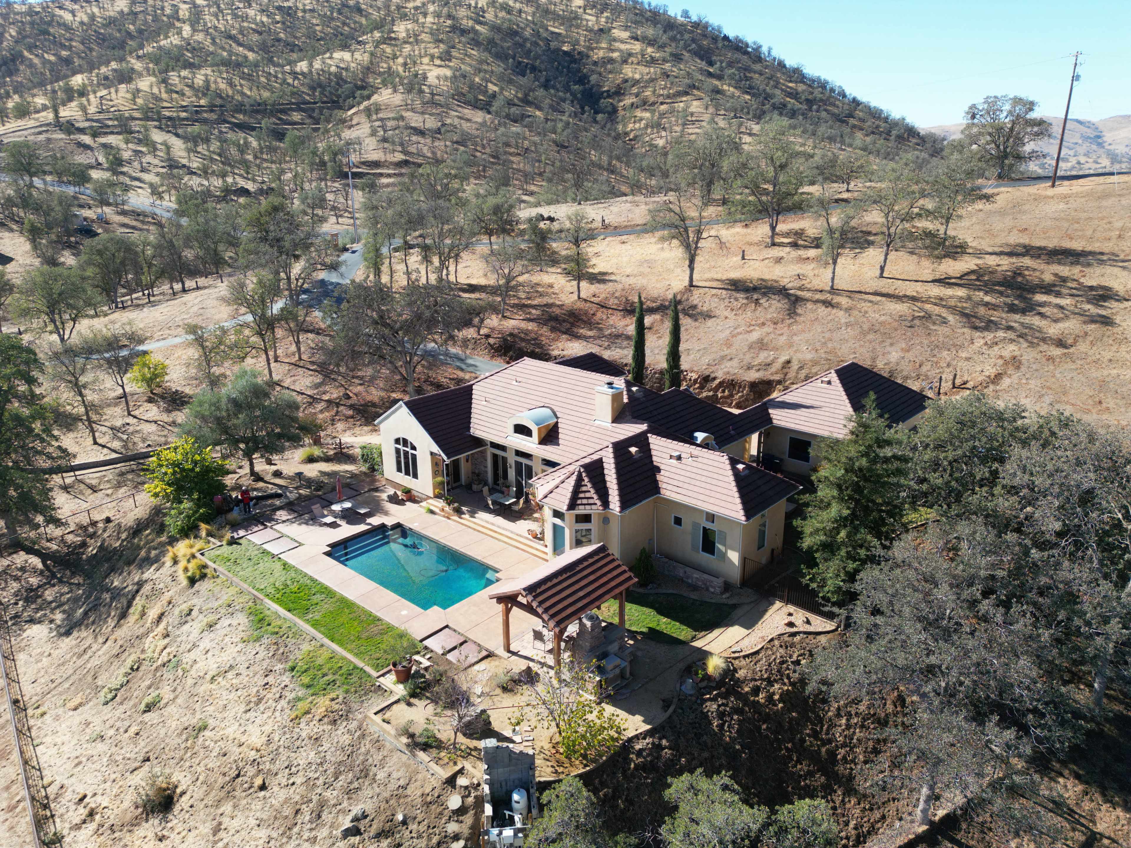 A spacious house with a pool is situated on a hillside surrounded by sparse trees and dry land.