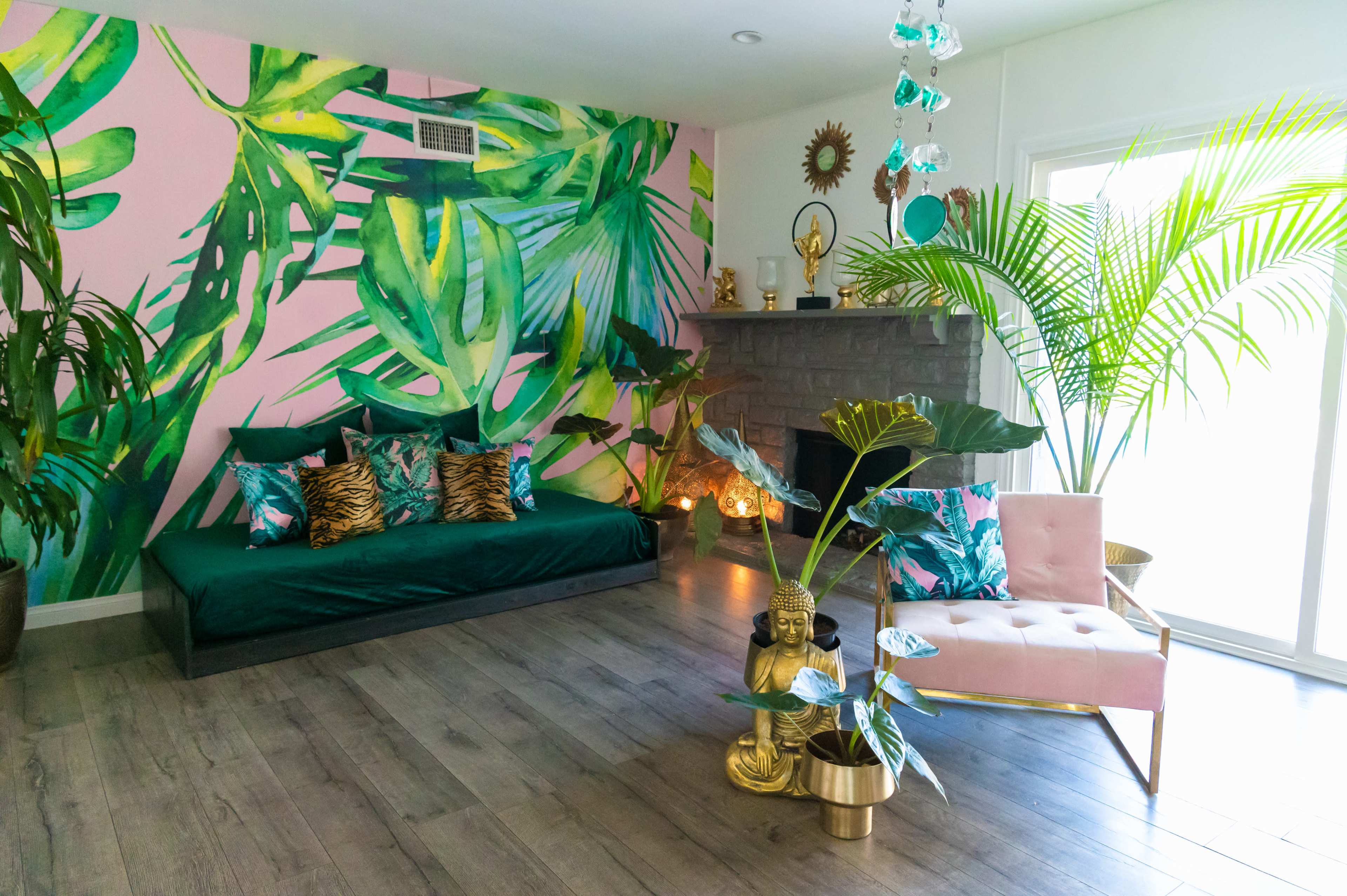 A vibrant living room with a large pink wall mural of tropical leaves, a green couch adorned with colorful pillows, and a pink chair beside a potted plant.