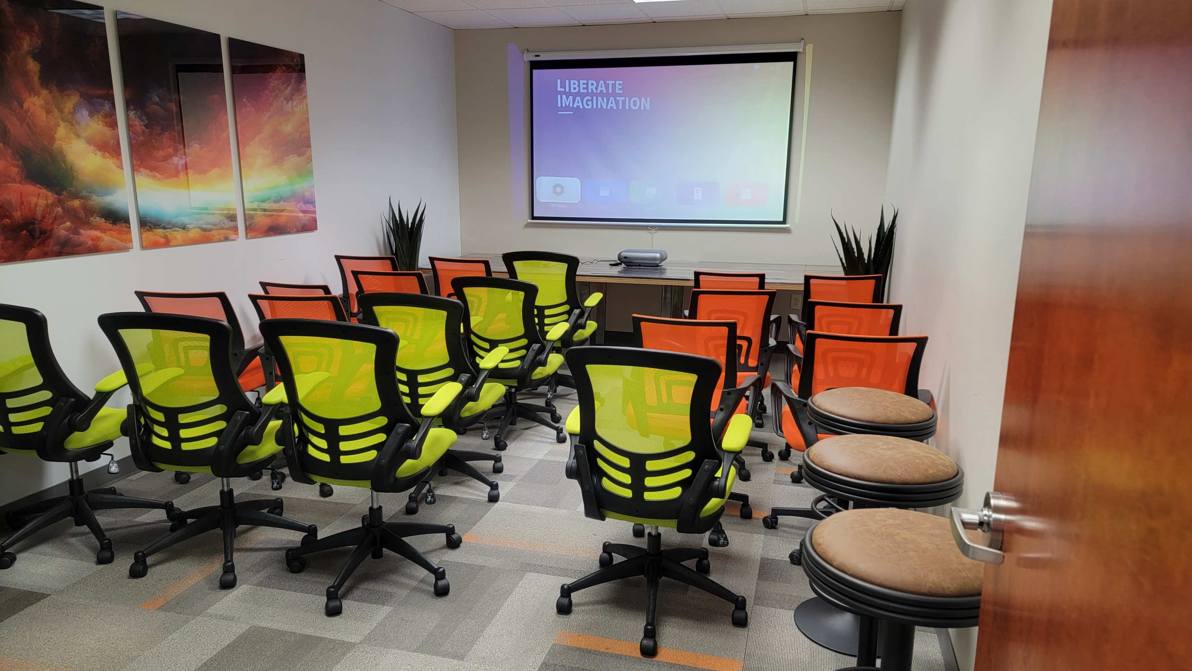 A conference room features rows of green and orange chairs facing a screen, with a table and potted plants in the background.