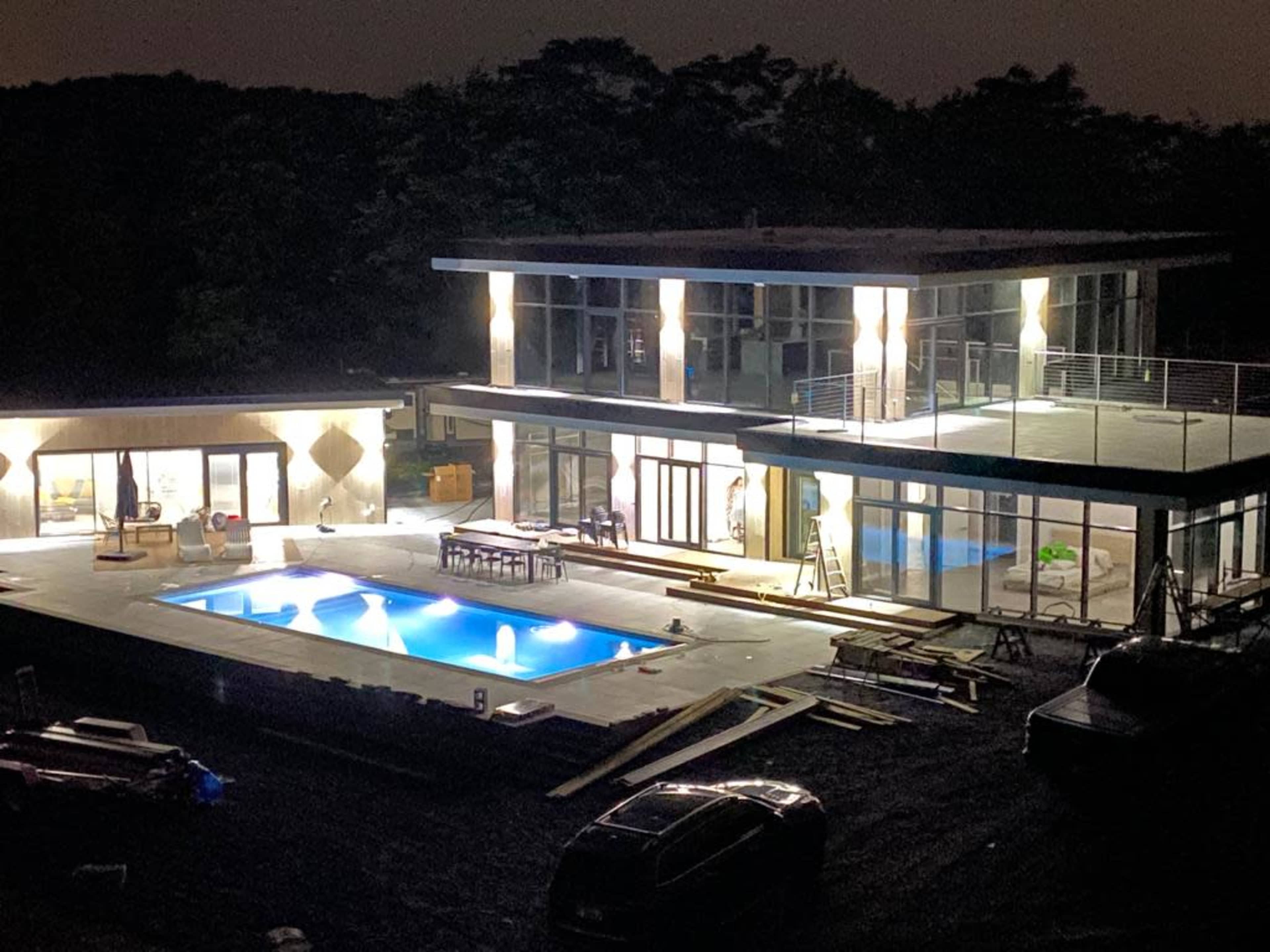 A modern two-story house with large windows and a swimming pool is illuminated at night, with construction materials and workers nearby.