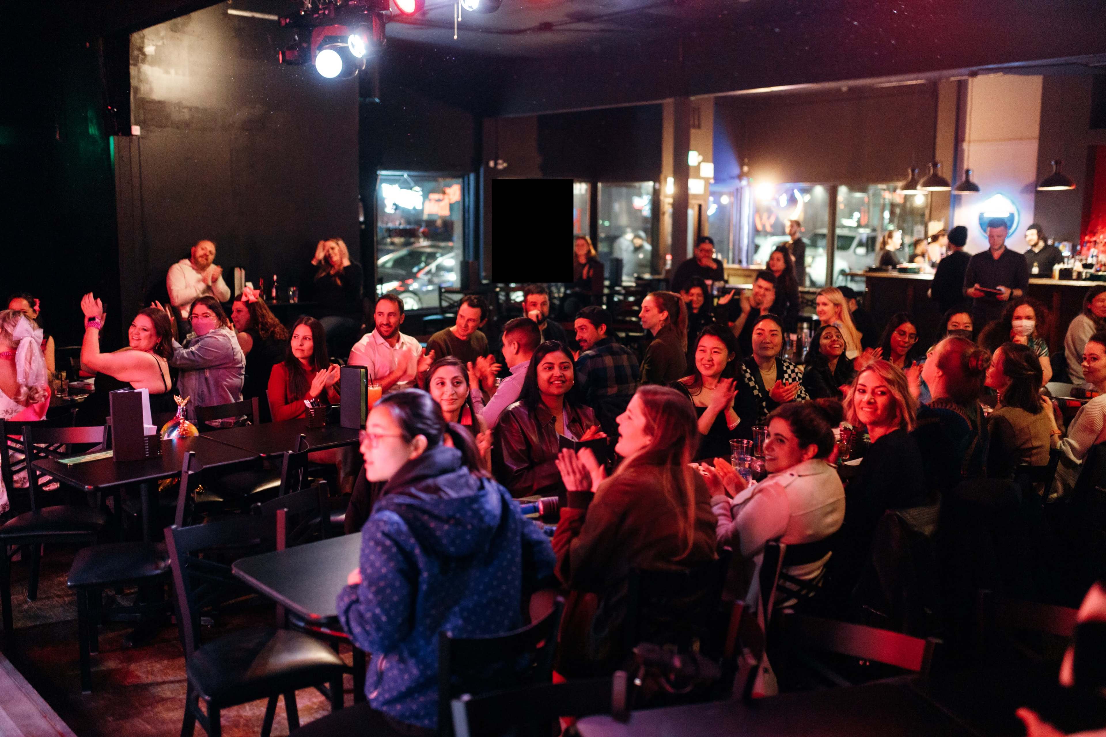 A crowd of people sits at tables in a dimly lit venue, some clapping and engaging with an event taking place on stage.