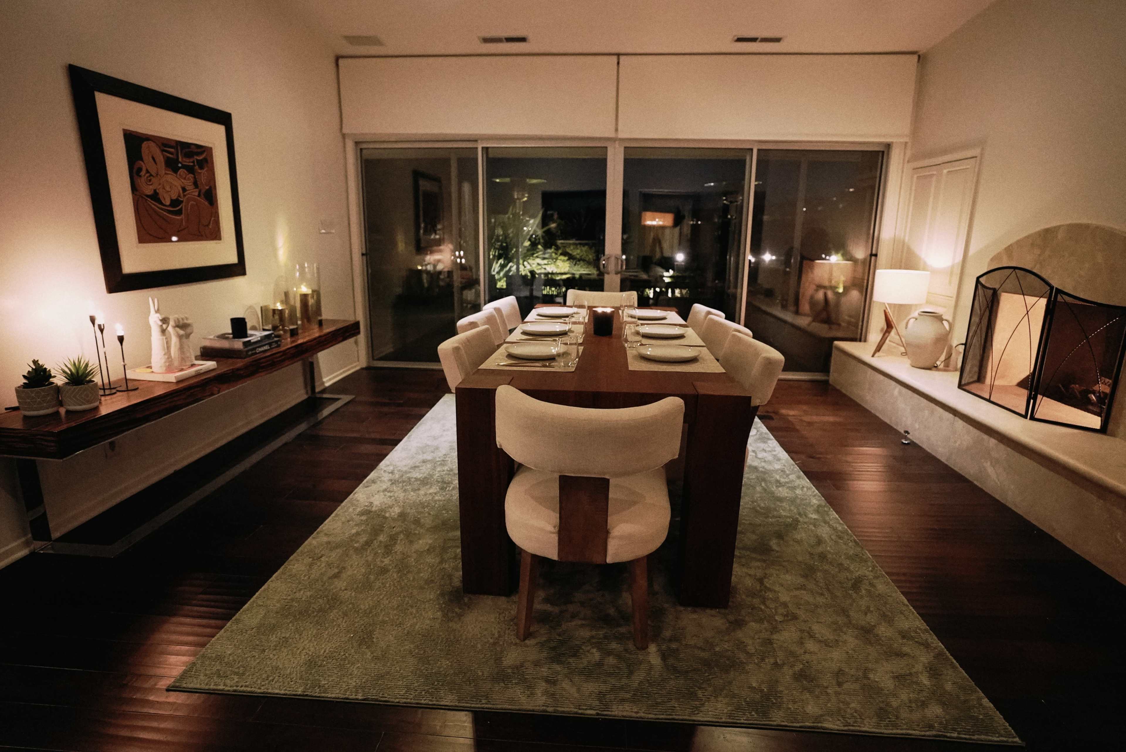 The image features a modern dining room with a large wooden table set for dinner, surrounded by eight chairs, and softly illuminated by ambient lighting.