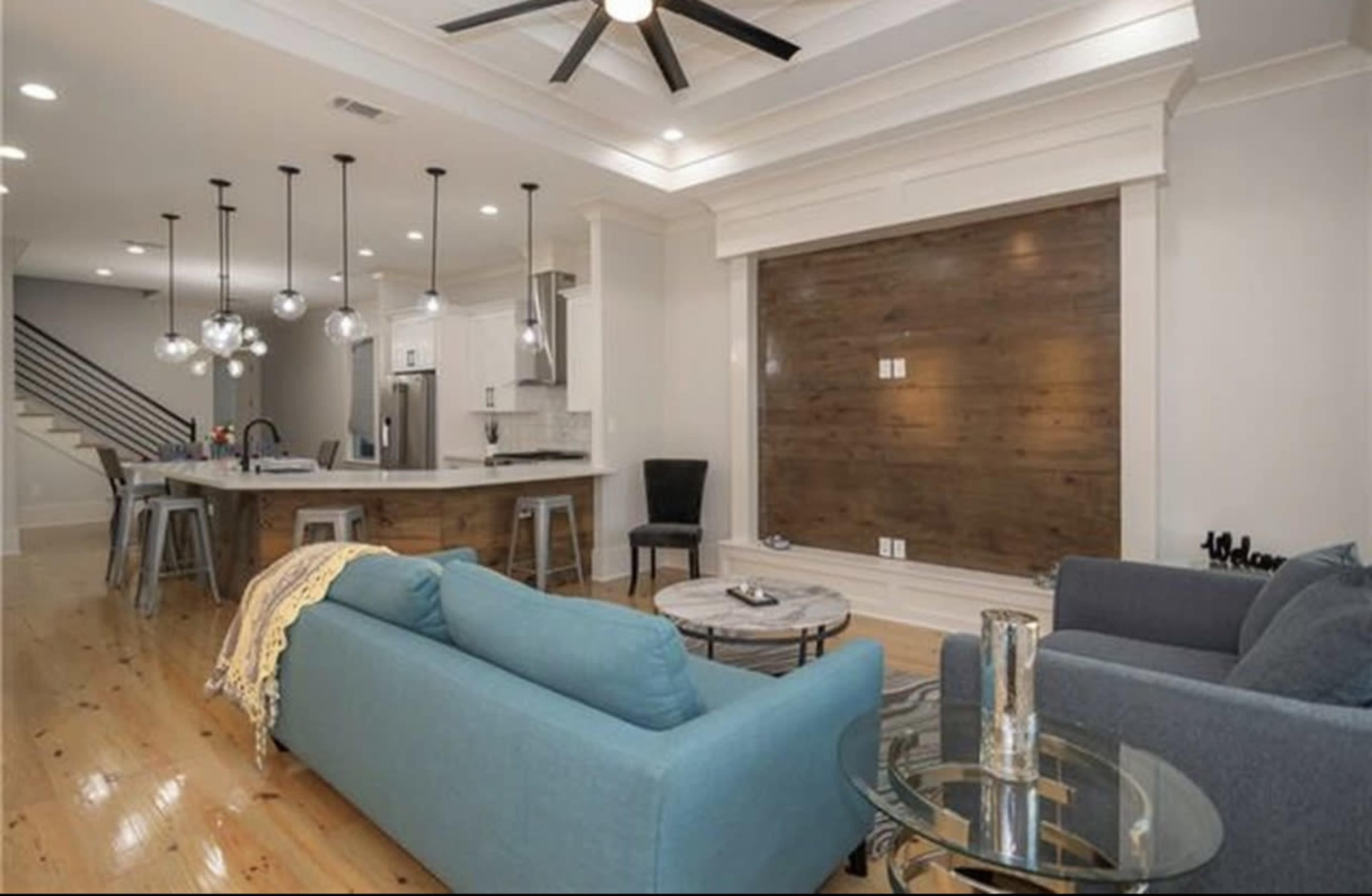 The image depicts a modern living room with a light-colored sofa, a dark blue armchair, a round coffee table, and a kitchen area featuring pendant lights and stainless steel appliances.