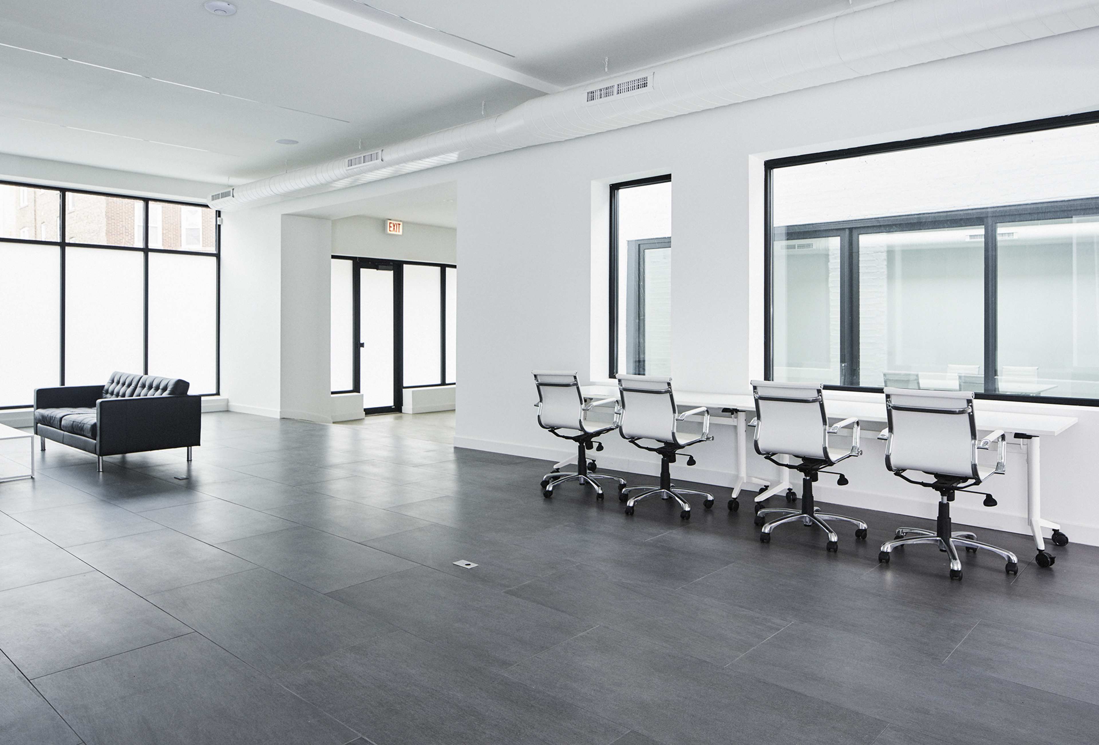 A modern, minimalist office space with a black couch and several office chairs arranged around a long desk, featuring large windows that allow natural light to enter.
