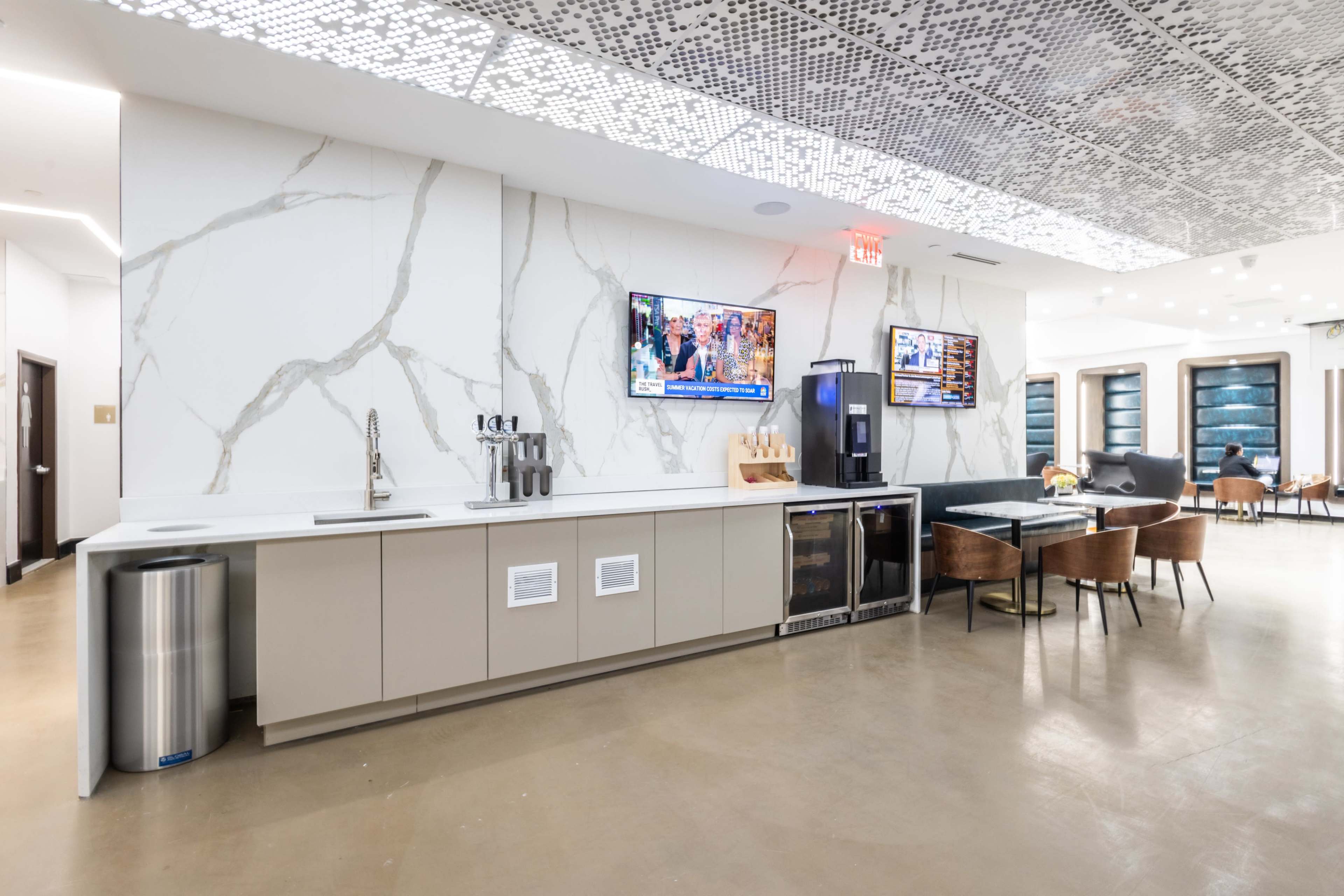 A modern kitchen with a large countertop, two televisions mounted on the wall, a beverage cooler, and a dining table with chairs.