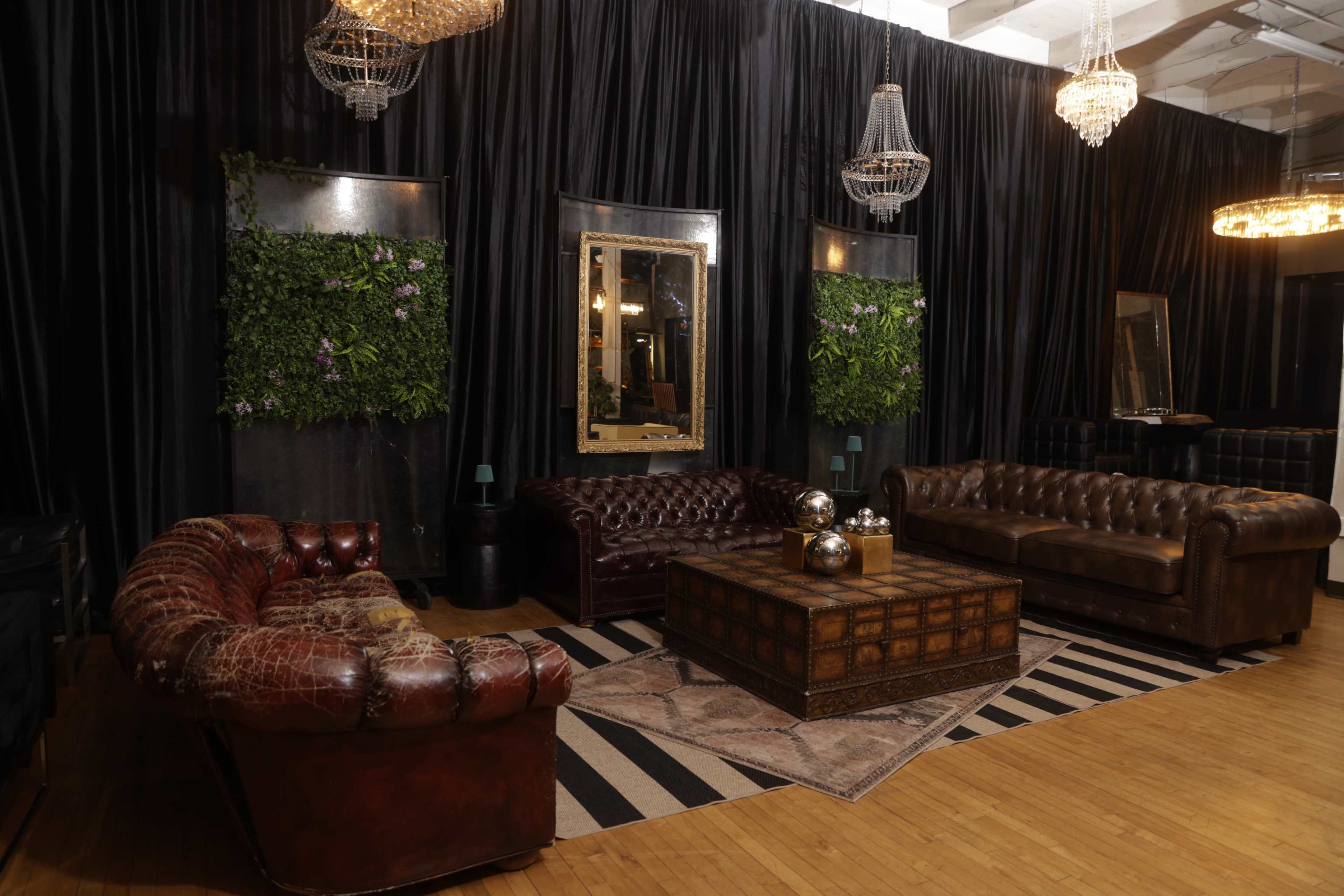 The image shows a stylish lounge area with dark walls, featuring two patterned leather sofas, a wooden coffee table, and decorative plants alongside ornate chandeliers.