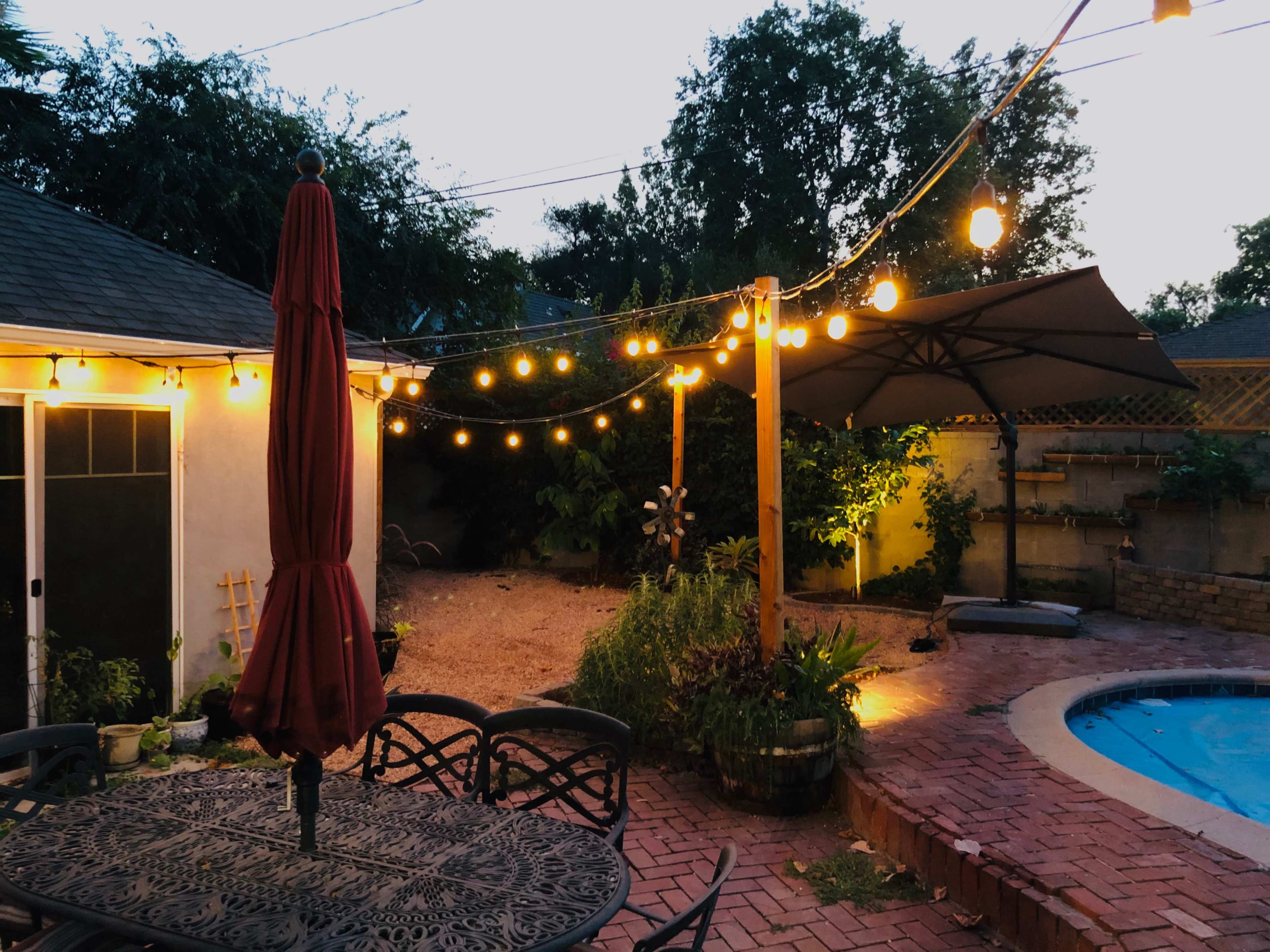 A backyard scene at dusk, featuring a patio table with a red umbrella, string lights overhead, a pool area, and greenery surrounding a small garden.