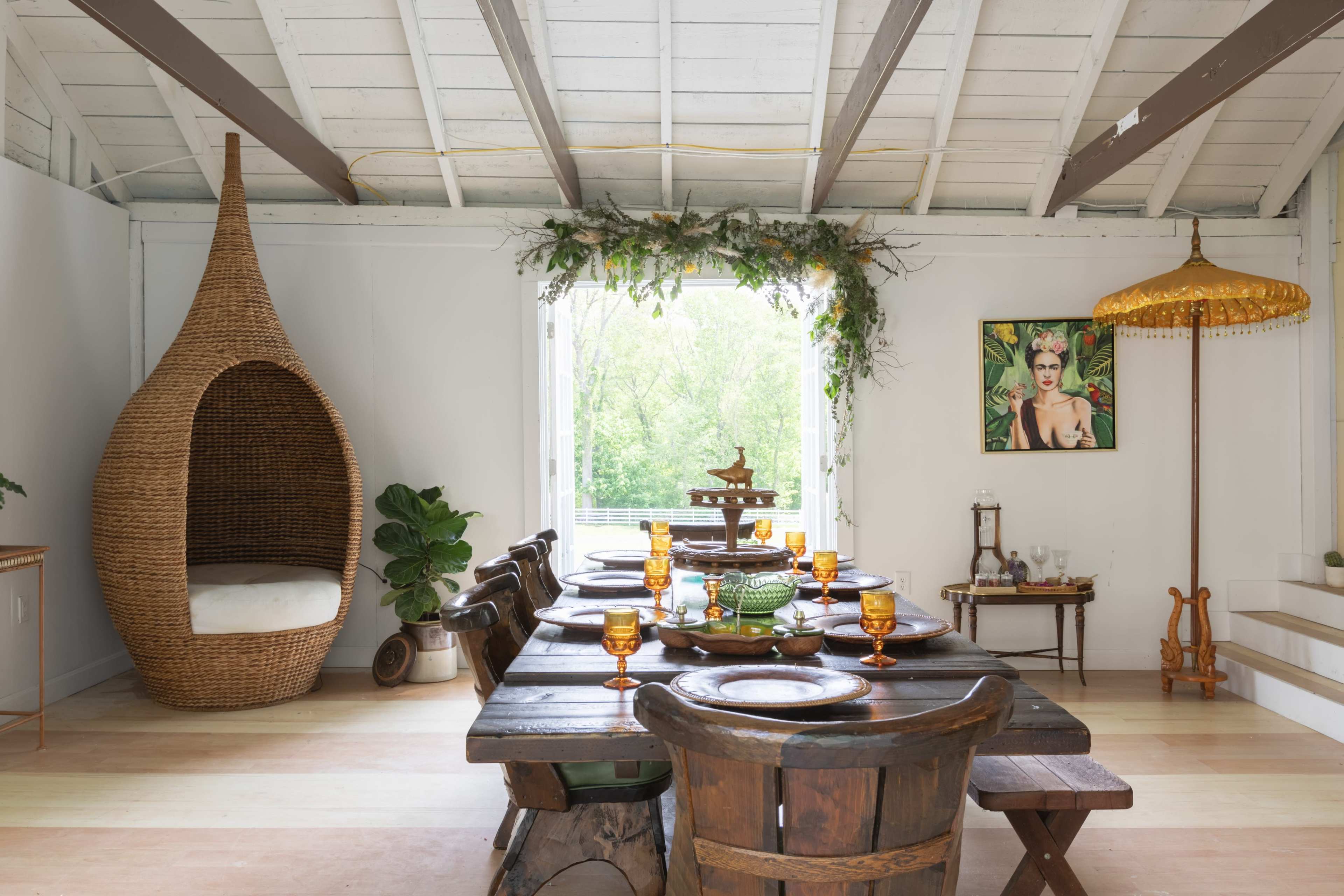 The image shows a dining area with a large wooden table set for a meal, surrounded by rustic chairs and featuring a woven hanging pod chair in one corner, alongside decorative plants and artwork on the walls.