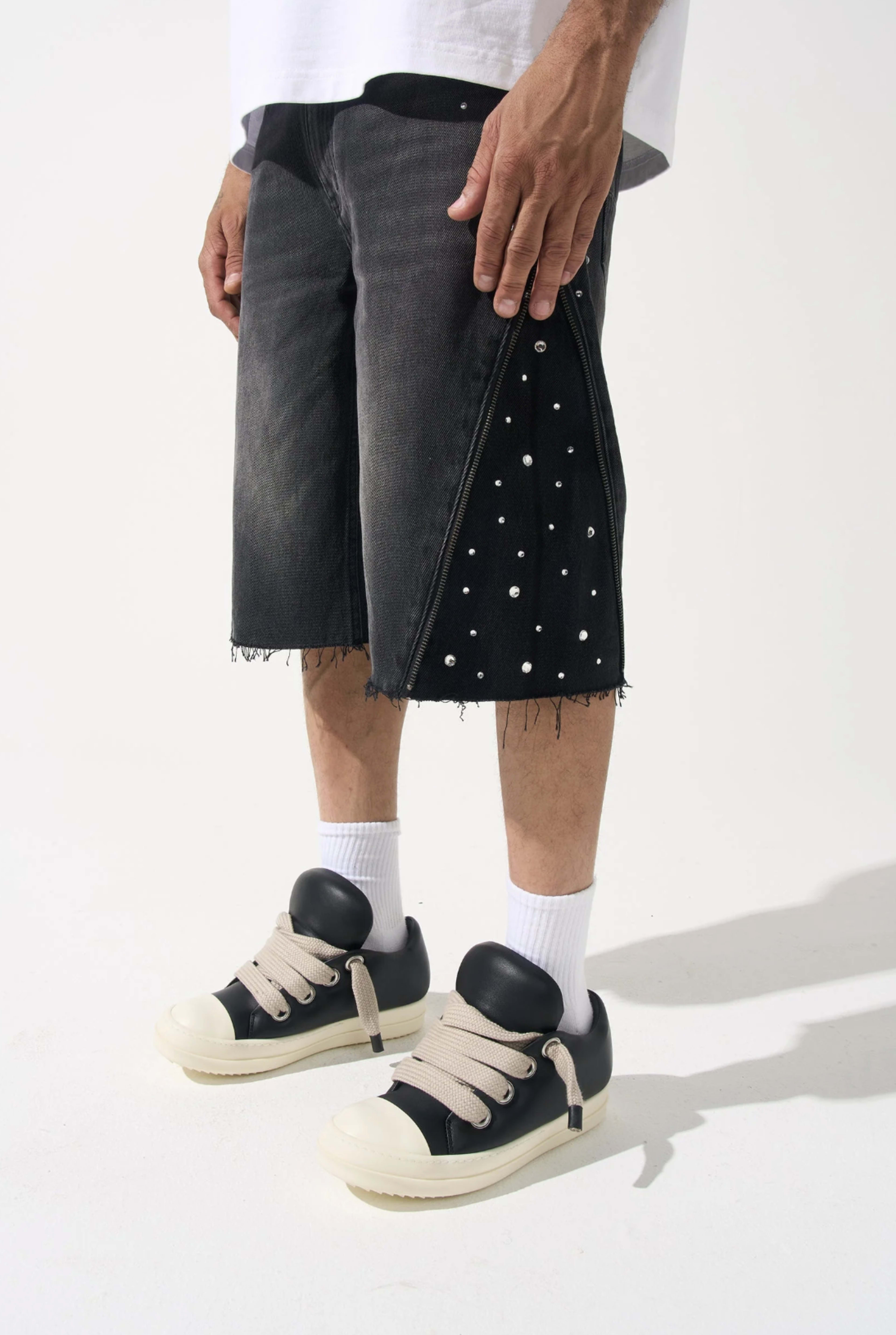 A person is wearing black denim shorts embellished with white studs and oversized black sneakers with light-colored soles.
