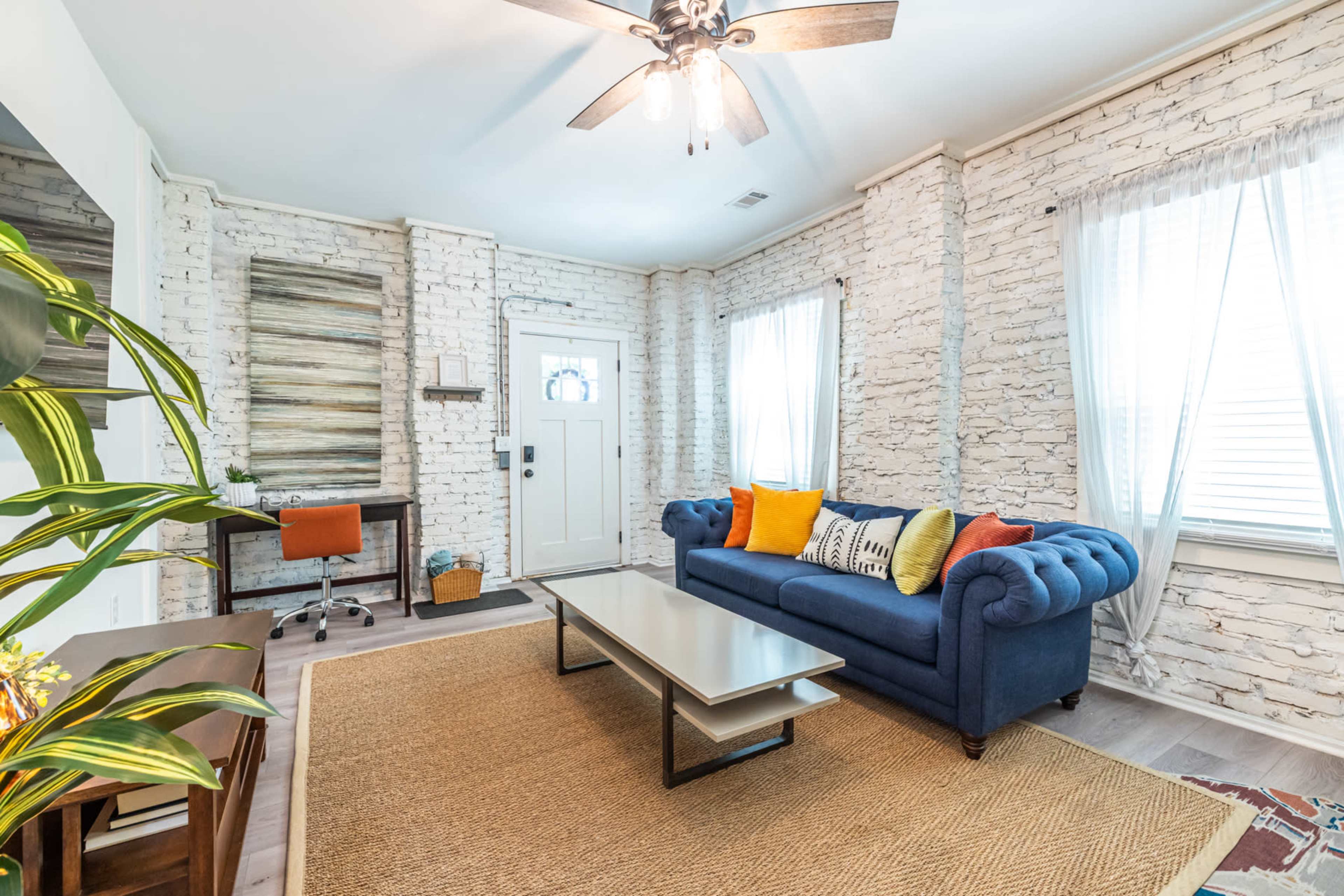 The image shows a cozy living room with white brick walls, a blue chesterfield sofa, a coffee table, and a small workspace with a desk and orange chair.