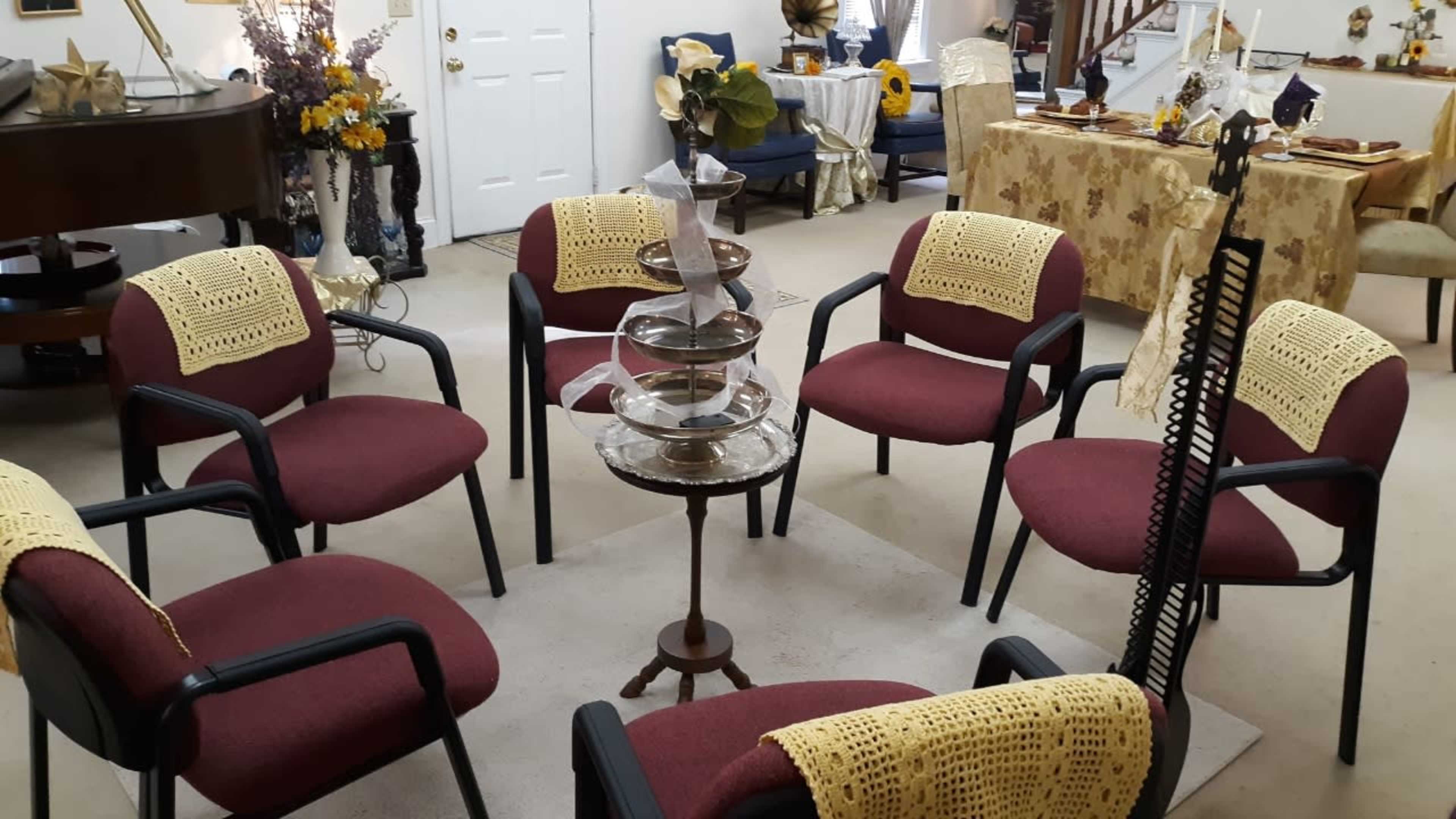 A circular arrangement of maroon chairs with crocheted covers around a small decorative table, set in a room with multiple tables and floral decorations.