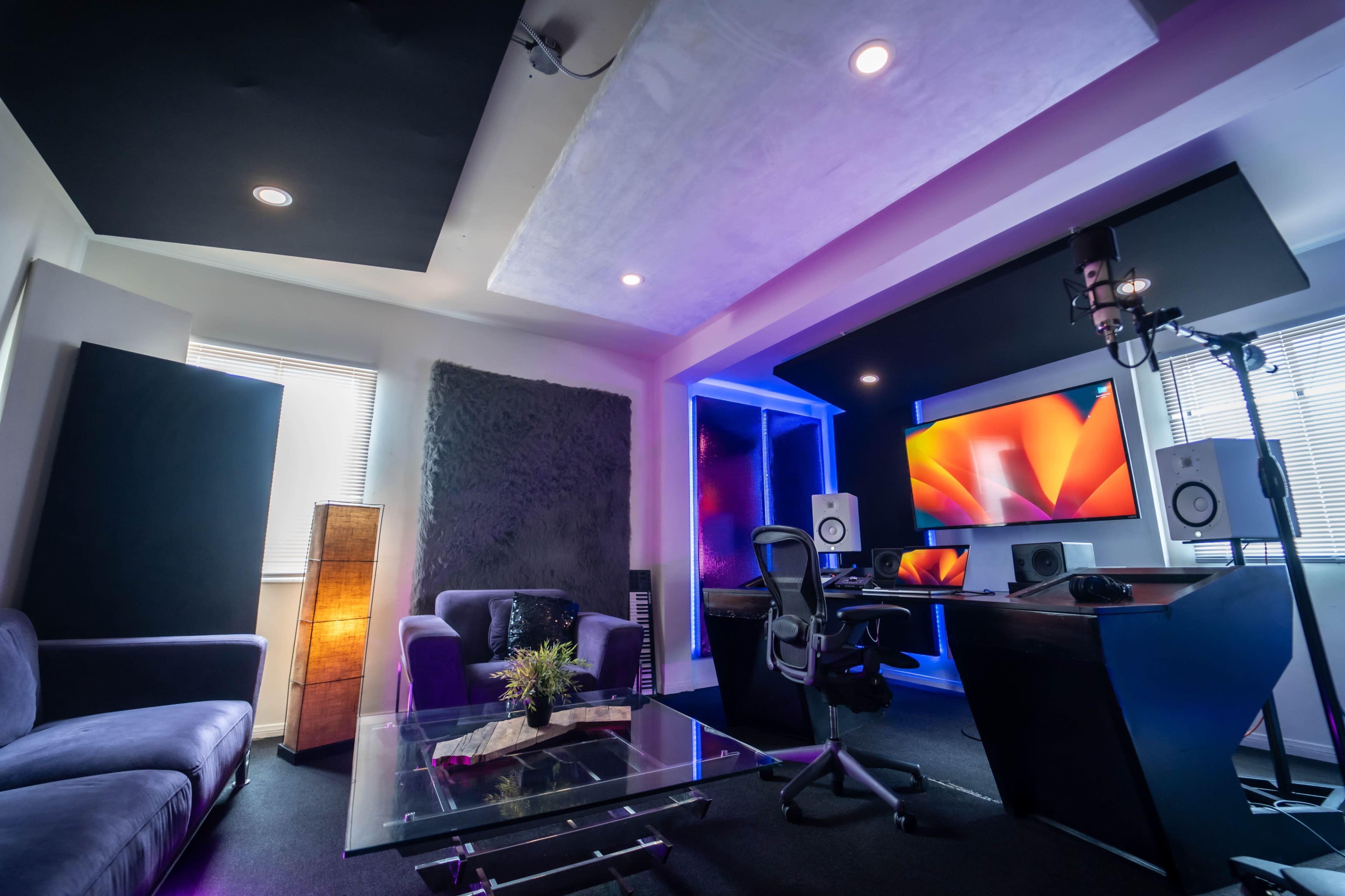 A modern recording studio with a desk, computer monitors, speakers, and a cozy seating area, featuring ambient lighting and soundproof panels.