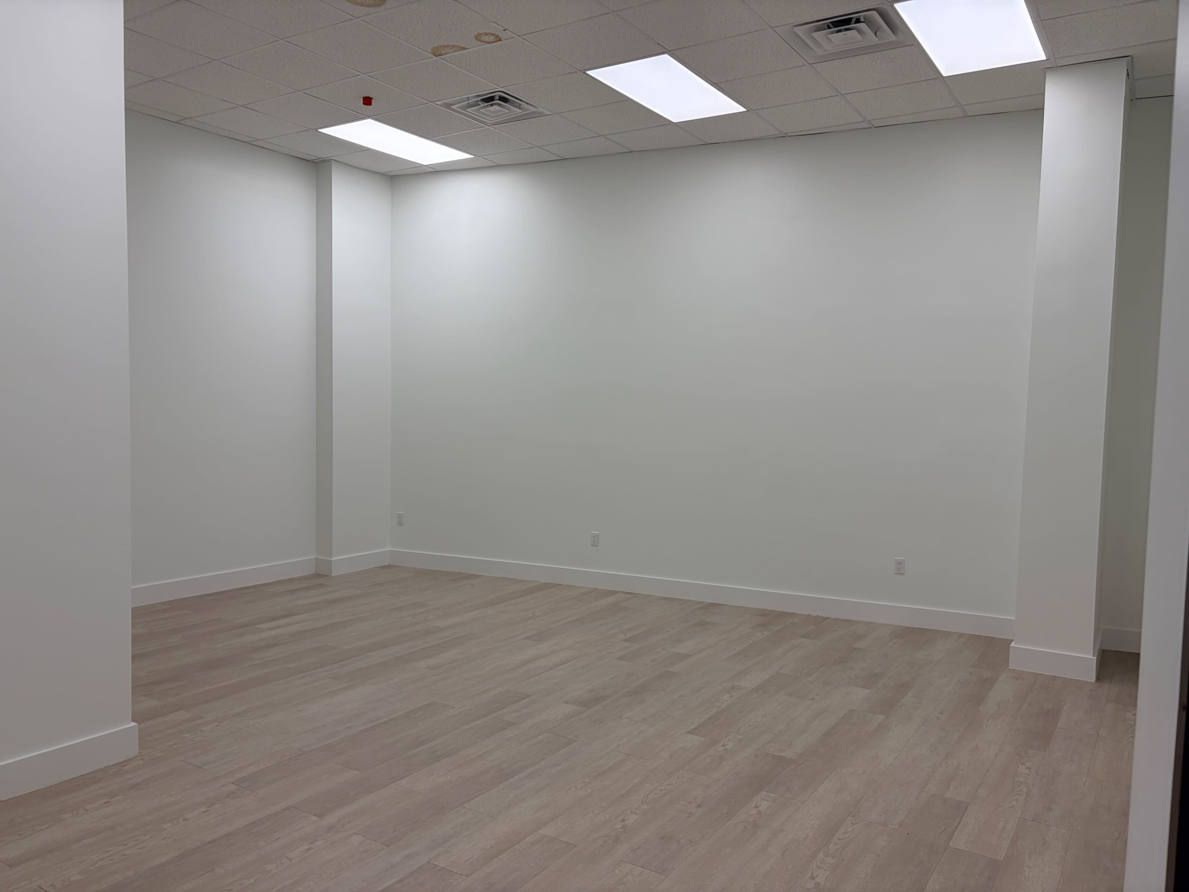The image shows an empty room with white walls and light-colored wood flooring.
