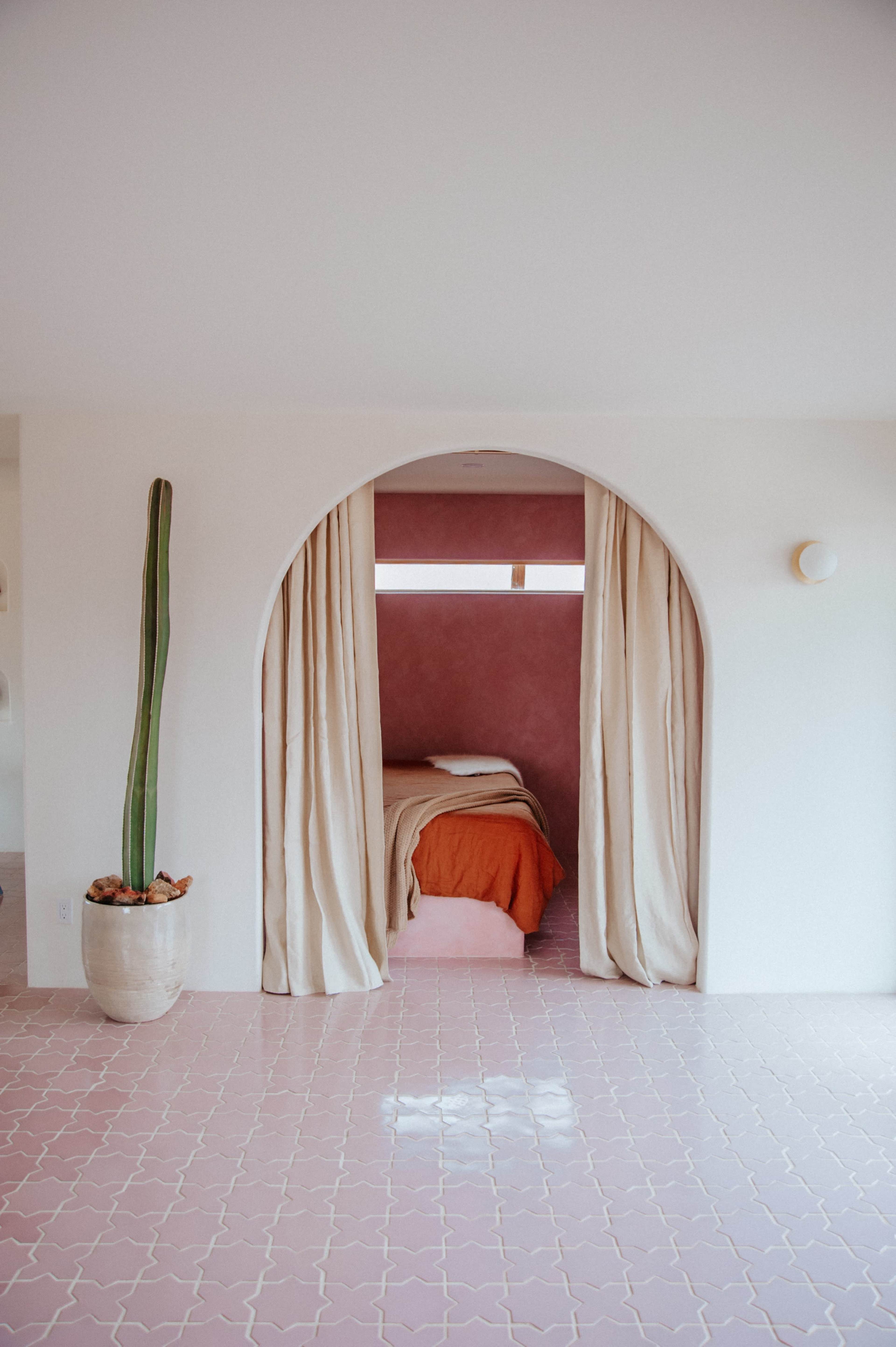 A minimalistic bedroom framed by a soft archway with beige curtains, pink walls, and a potted cactus in a decorative pot.