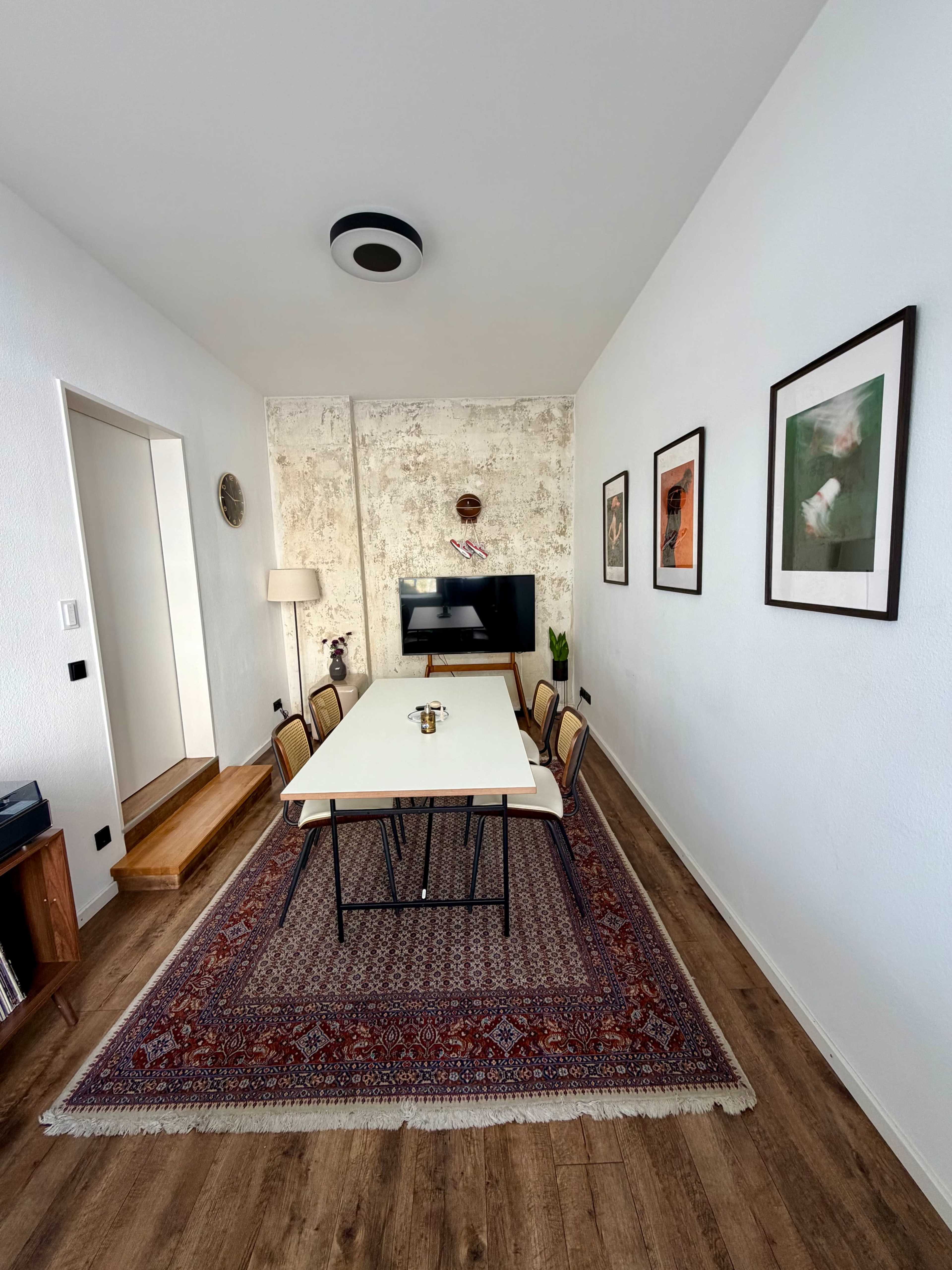 The image shows a narrow dining room with a long table surrounded by chairs, a television on the wall, and framed artwork, with a rug covering the wooden floor.