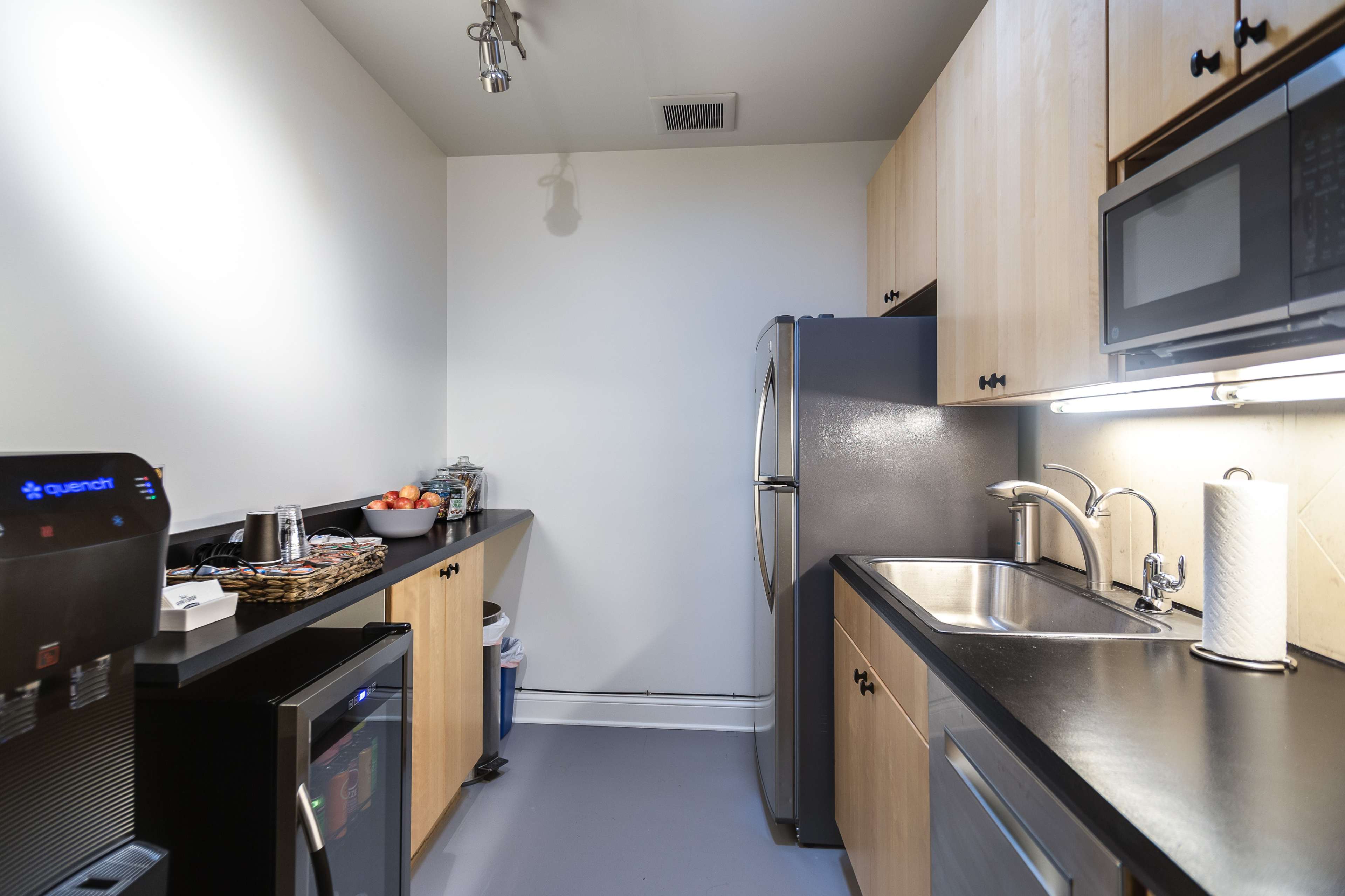A small kitchen features black countertops, wooden cabinetry, a stainless steel refrigerator, a microwave, and an under-counter fridge.