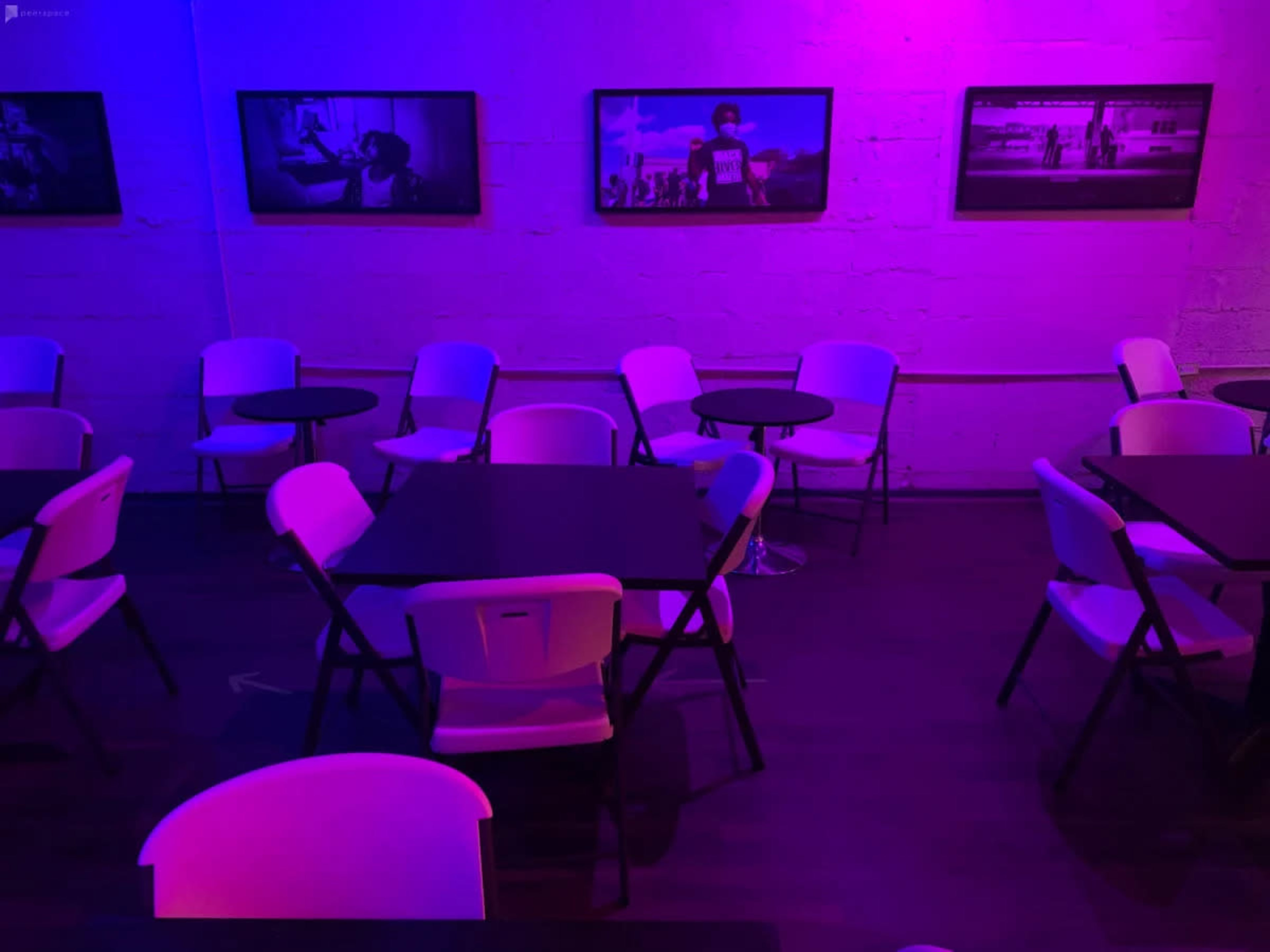 A dimly lit room features several tables and chairs, with purple lighting and black-and-white photographs hanging on the walls.